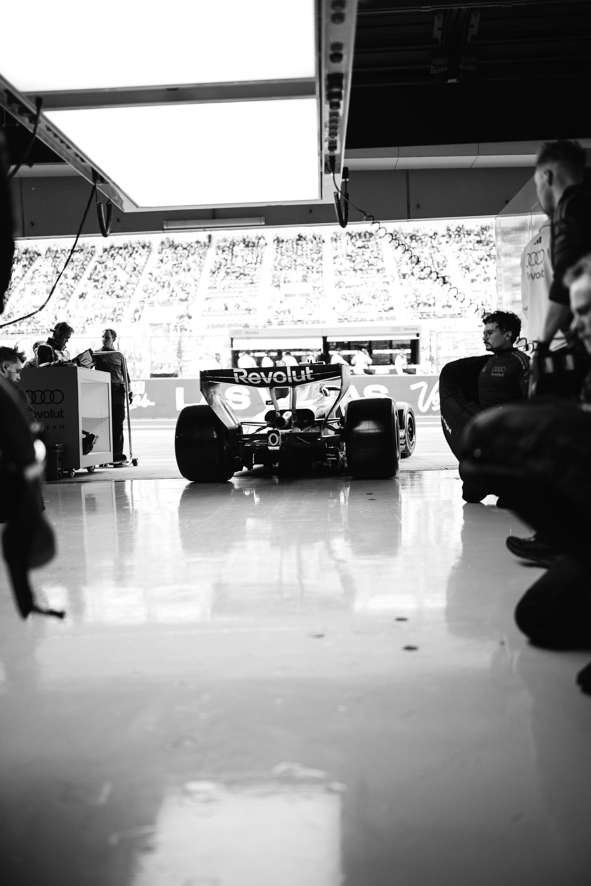 Black and white view of the Audi Revolut F1® Team car from behind in the garage, framed by team members and the bright pit lane beyond.
