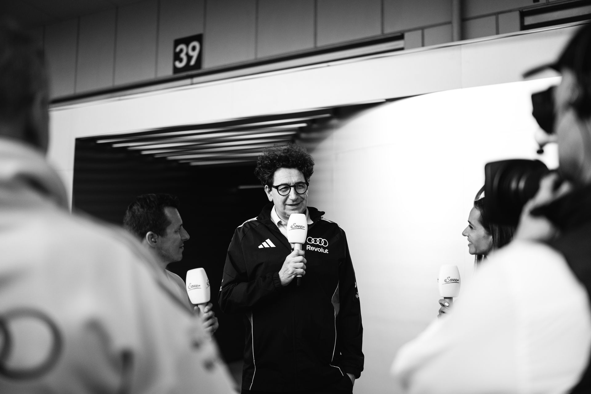 Black and white image of Mattia Binotto being interviewed between two presenters in the paddock.