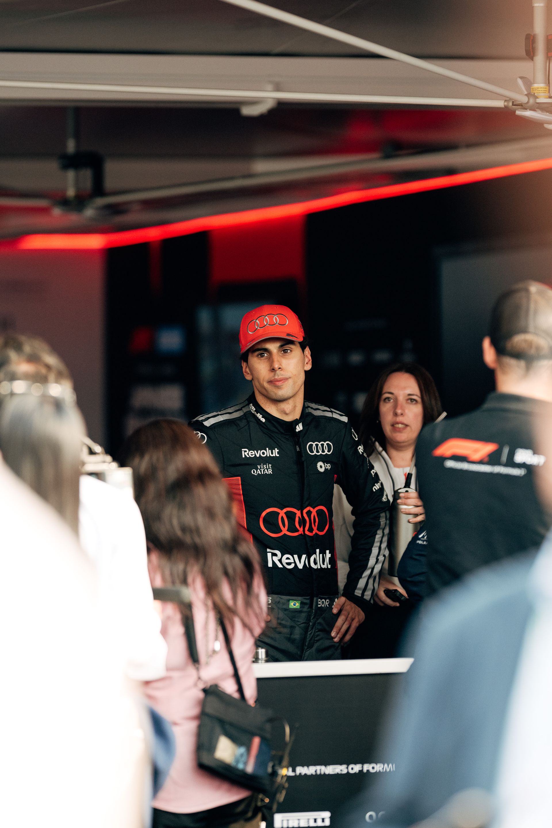 Gabriel Bortoleto stands in the Audi Revolut F1® Team garage wearing his race suit and red cap while people move around him.