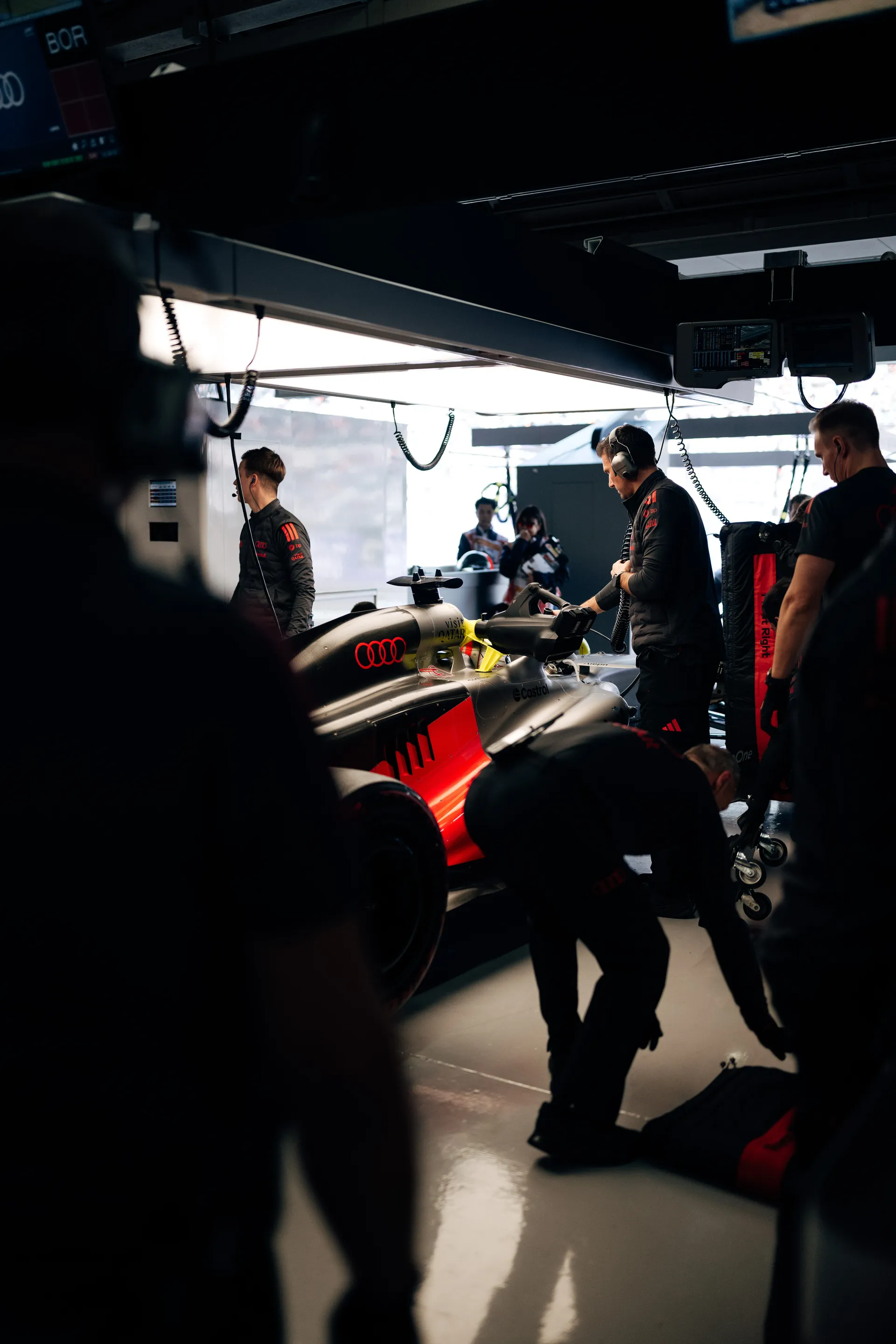 Inside the Audi Revolut F1® Team garage, mechanics work around the car under dark lighting and red illuminated bodywork accents.