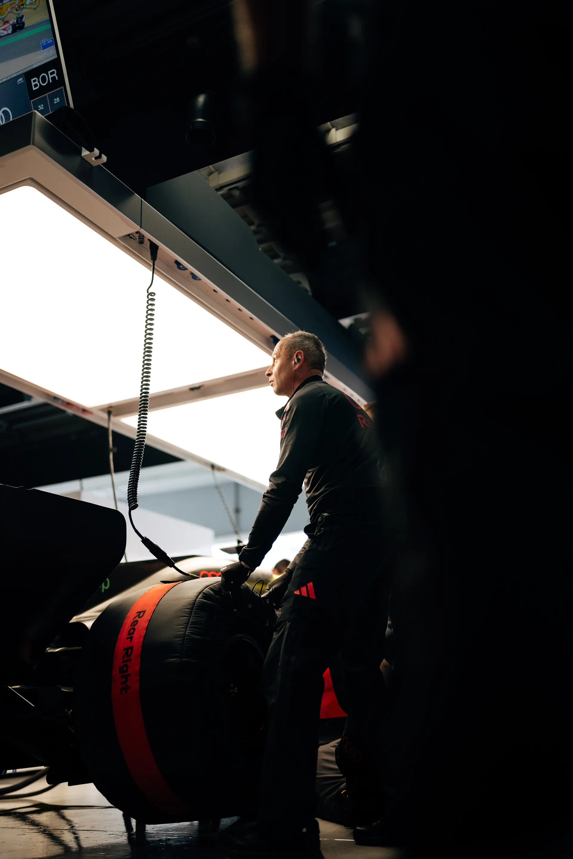A team mechanic stands beside a covered tire under the garage light panels while the Audi Revolut F1® Team car is prepared in the background.