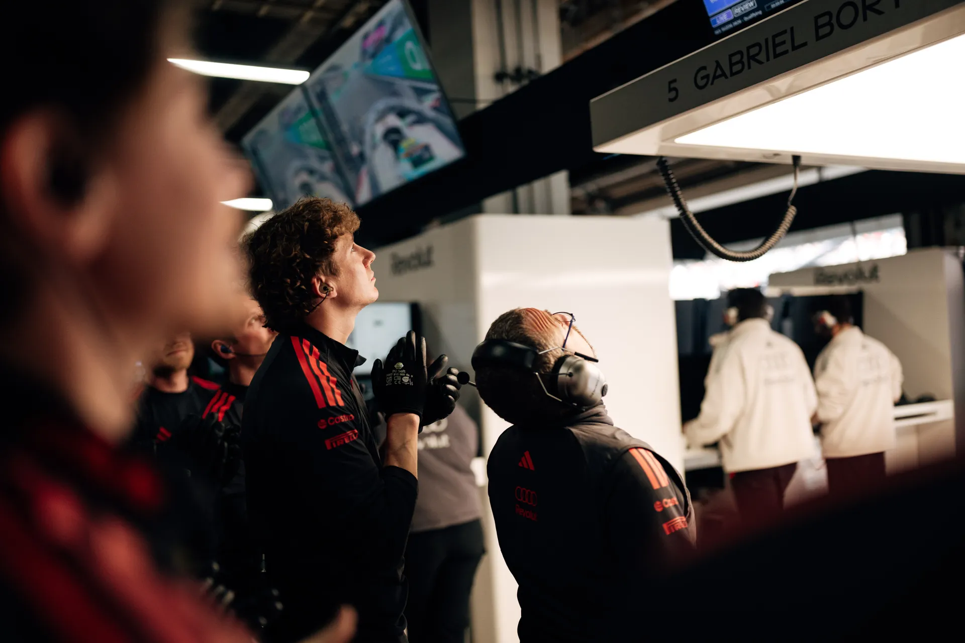 Several Audi Revolut F1® Team crew members gather beneath the Gabriel Bortoleto garage sign, watching overhead screens during a garage session.
