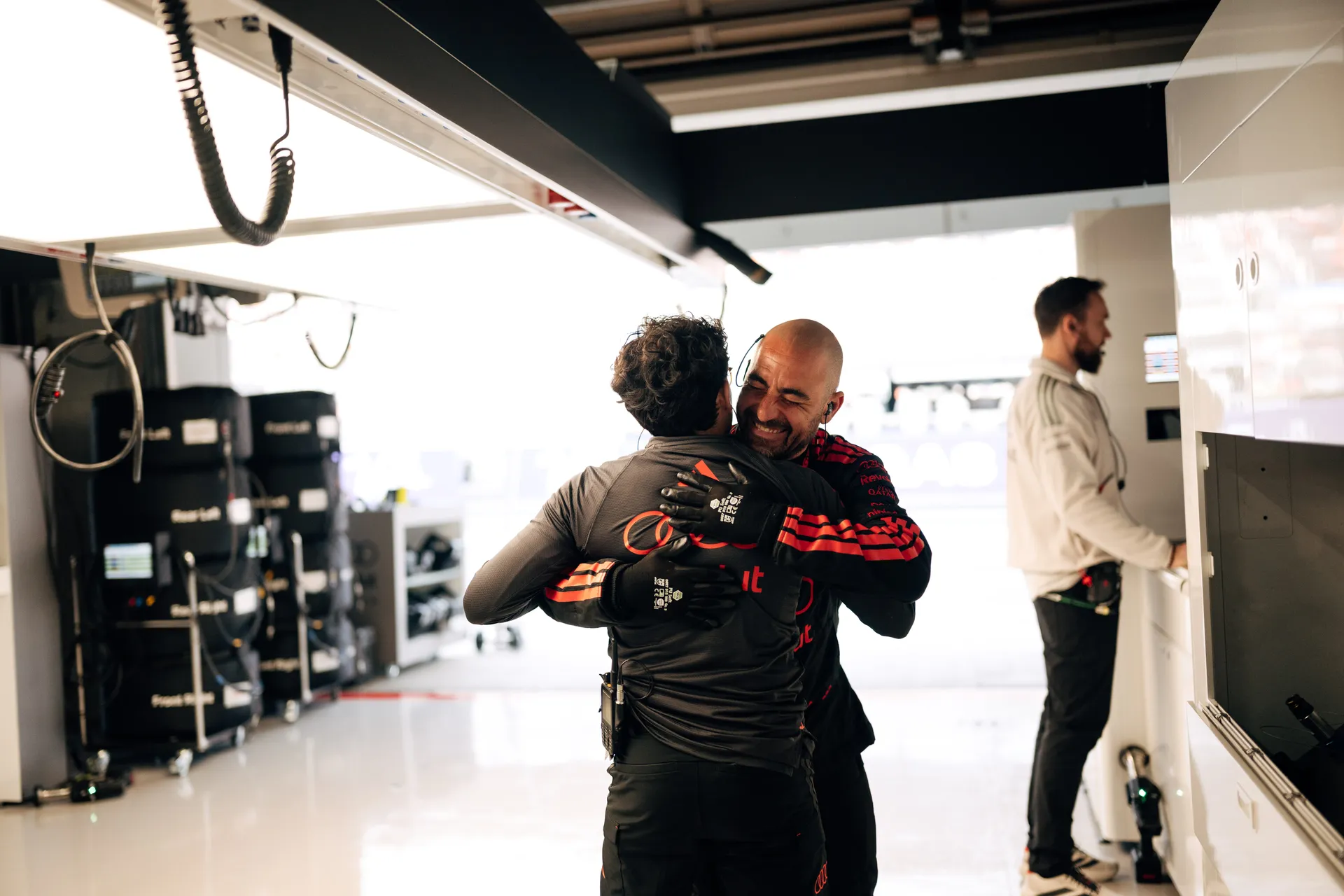 Two Audi Revolut F1® Team crew members share a hug in the garage while another team member works at a cabinet in the background.
