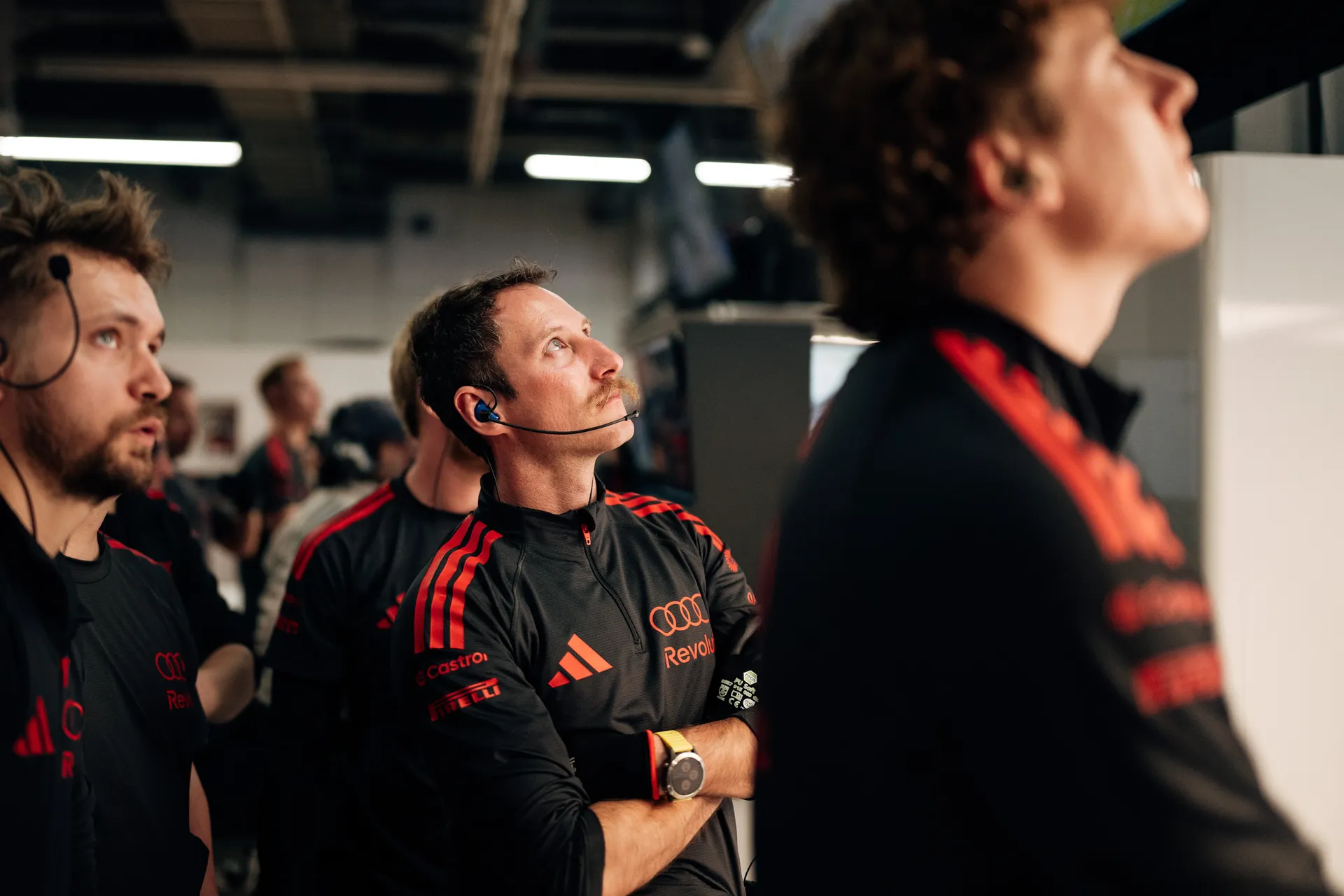 Audi Revolut F1® Team crew members in the garage look up toward screens, listening through headsets during an active session.