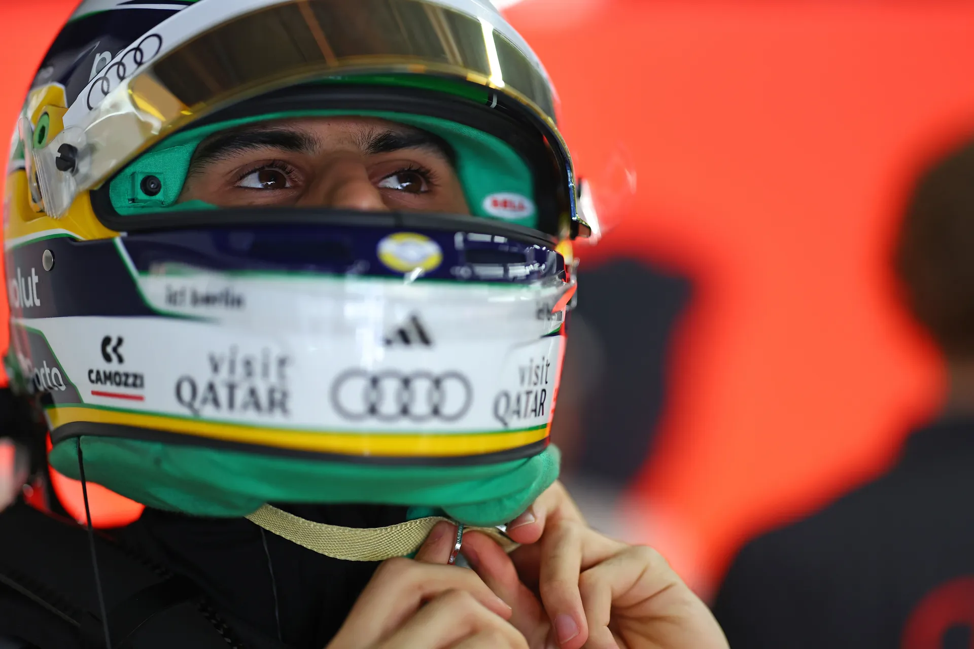 Close-up of Gabriel Bortoleto fastening his helmet strap in the Audi Revolut F1® Team garage, with a red-lit background behind him.