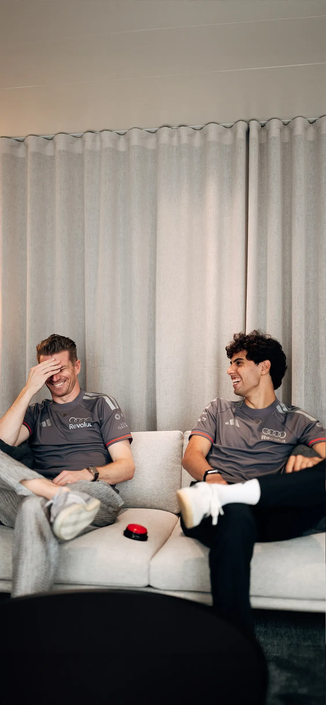 Nico Hulkenberg and Gabriel Bortoleto sitting on a couch in a relaxed indoor setting, casually talking and laughing, representing the lifestyle side of the Audi Revolut F1® Team away from the track