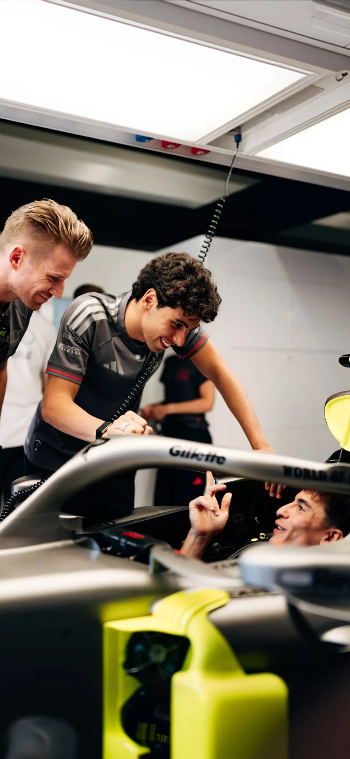 Marc Márquez sits in the cockpit of the Audi Revolut F1® Team car Audi R26 inside the garage while Nico Hulkenberg and Gabriel Bortoleto stand beside the car observing and discussing