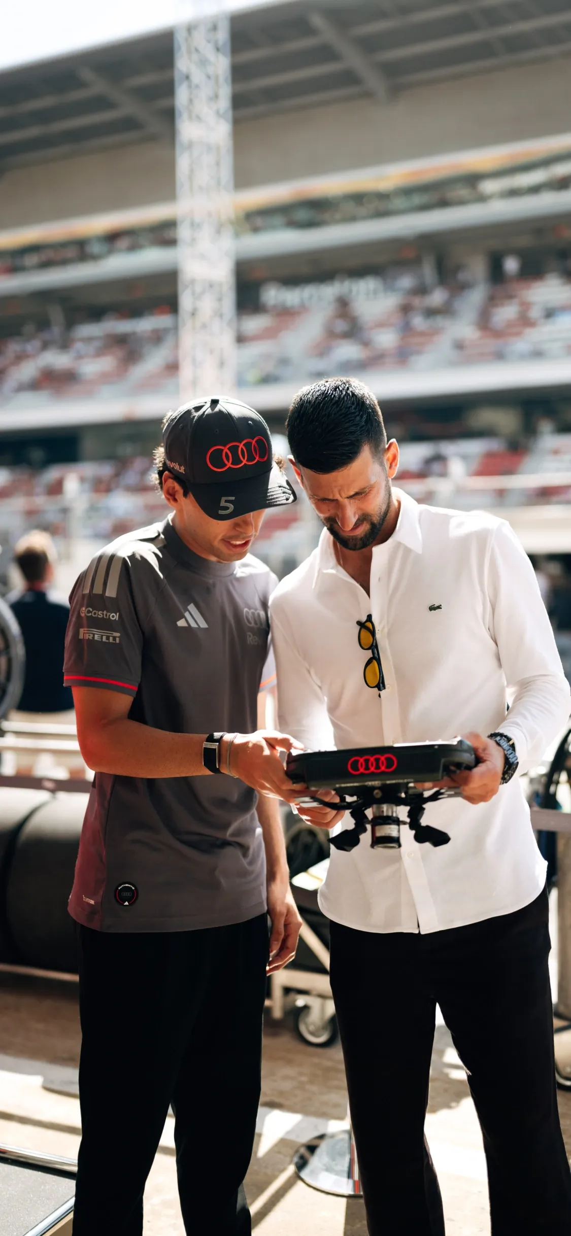 Gabriel Bortoleto and Novak Djokovic standing trackside reviewing data on a handheld device during a race weekend for the Audi Revolut F1® Team