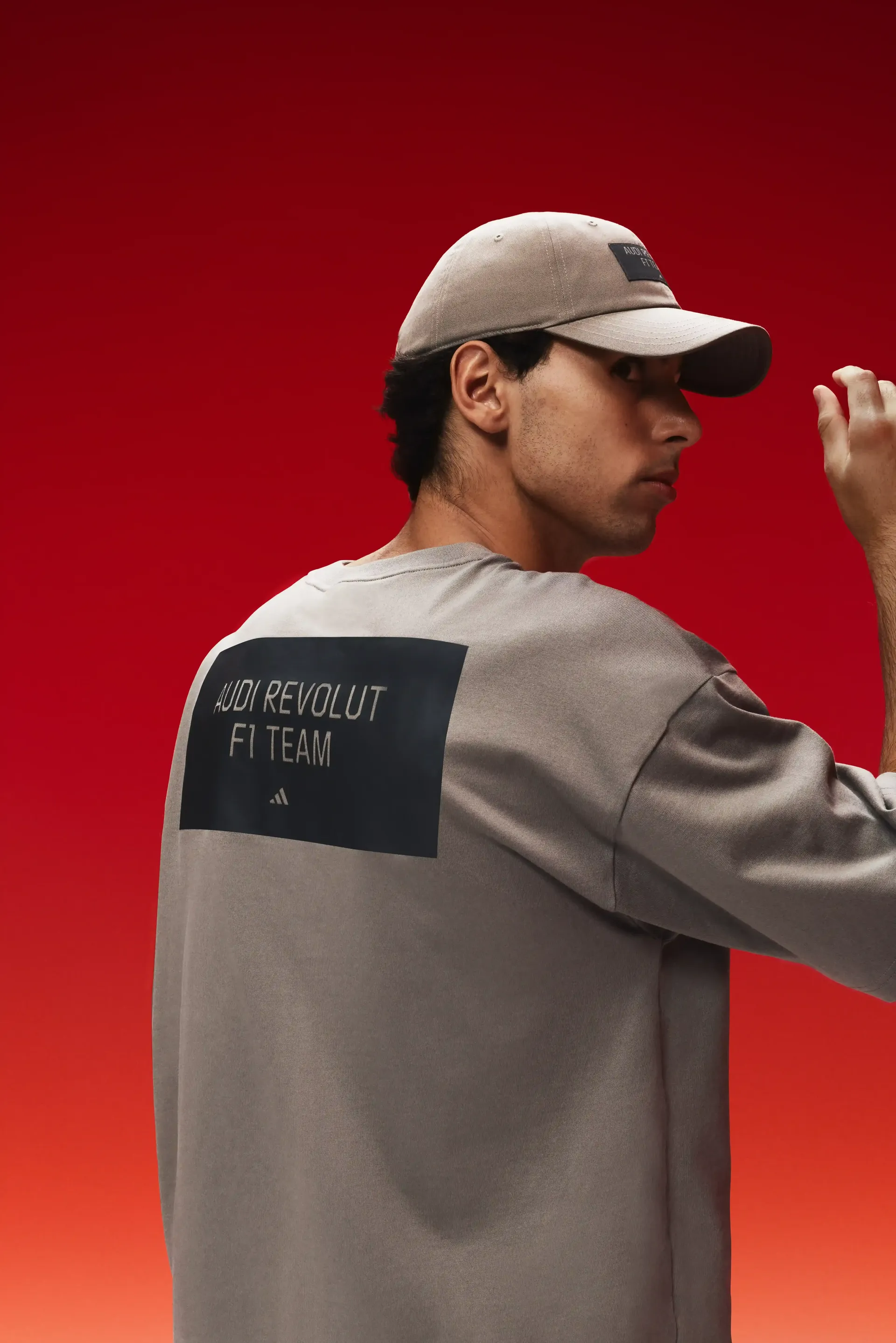 Formula 1 driver Gabriel Bortoleto seen from behind wearing a light-brown team shirt, visible back branding, cap, red studio background, focus on team identity.