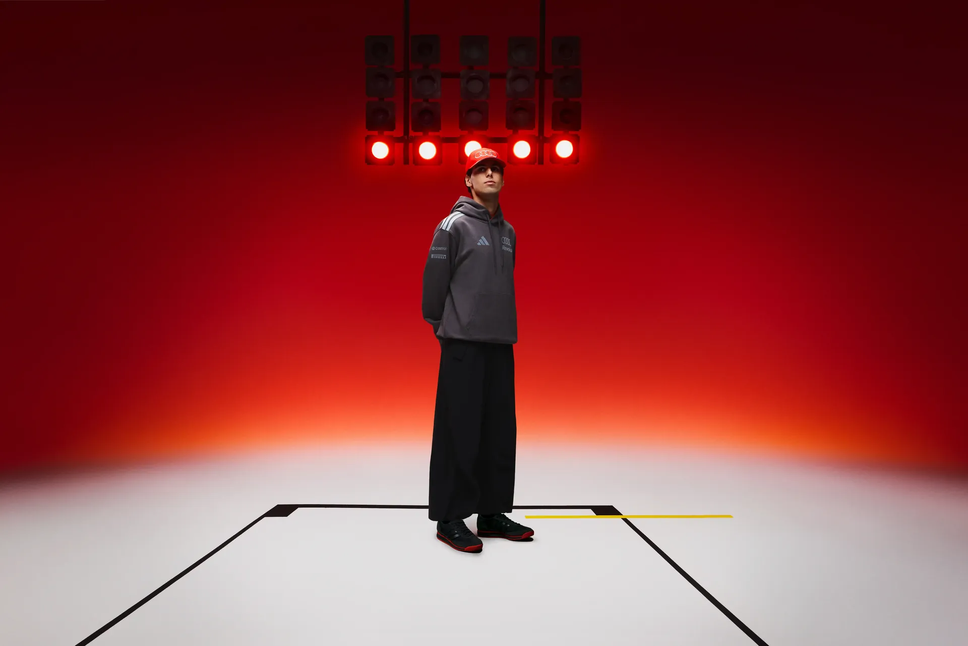 Full-body shot of Formula 1 driver Gabriel Bortoleto Revolut Audi F1 team alone in studio white floor with racing line graphics red light gradient background grey hoodie black pants