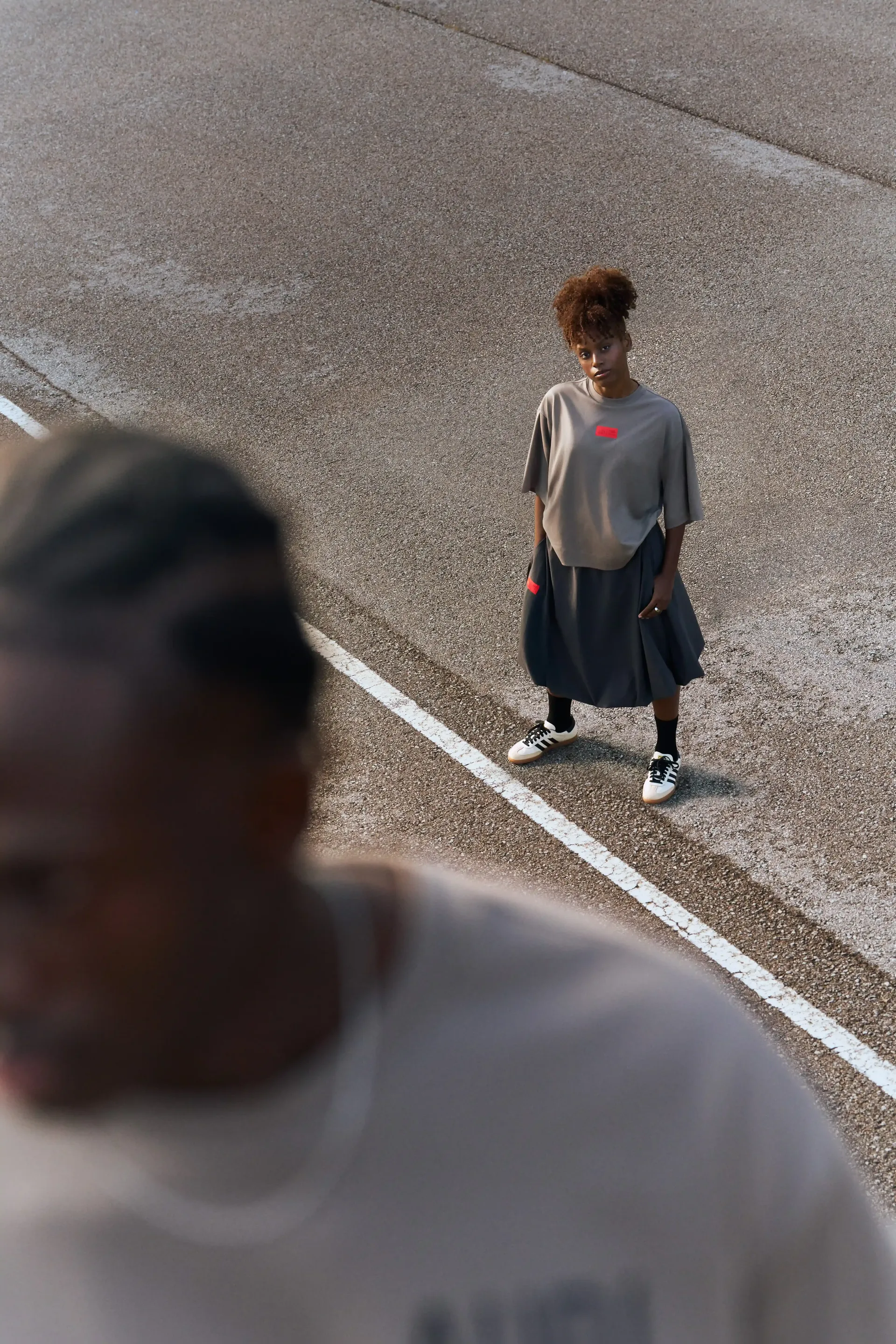 Two models, the one in front shown in closeup, standing on a concrete road, wearing gray-brown shirts and a dark skirt, relaxed posture with a modern streetwear look.