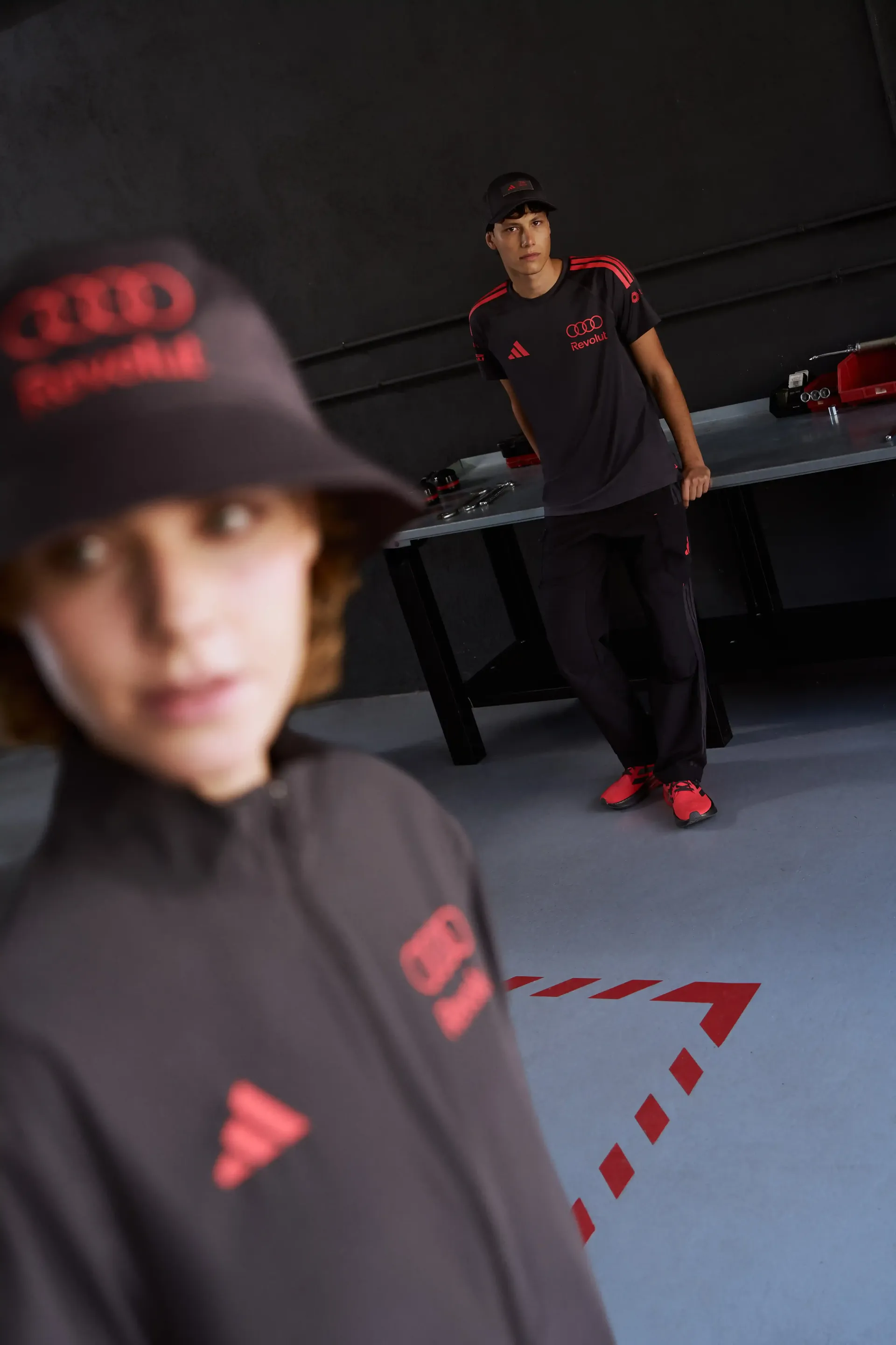 Close-up of a model wearing cap focus on facial expression dark outfit motorsport-inspired style, Adidas mechanic wear, red shoes, Formel 1 Audi Revolut Teamwear