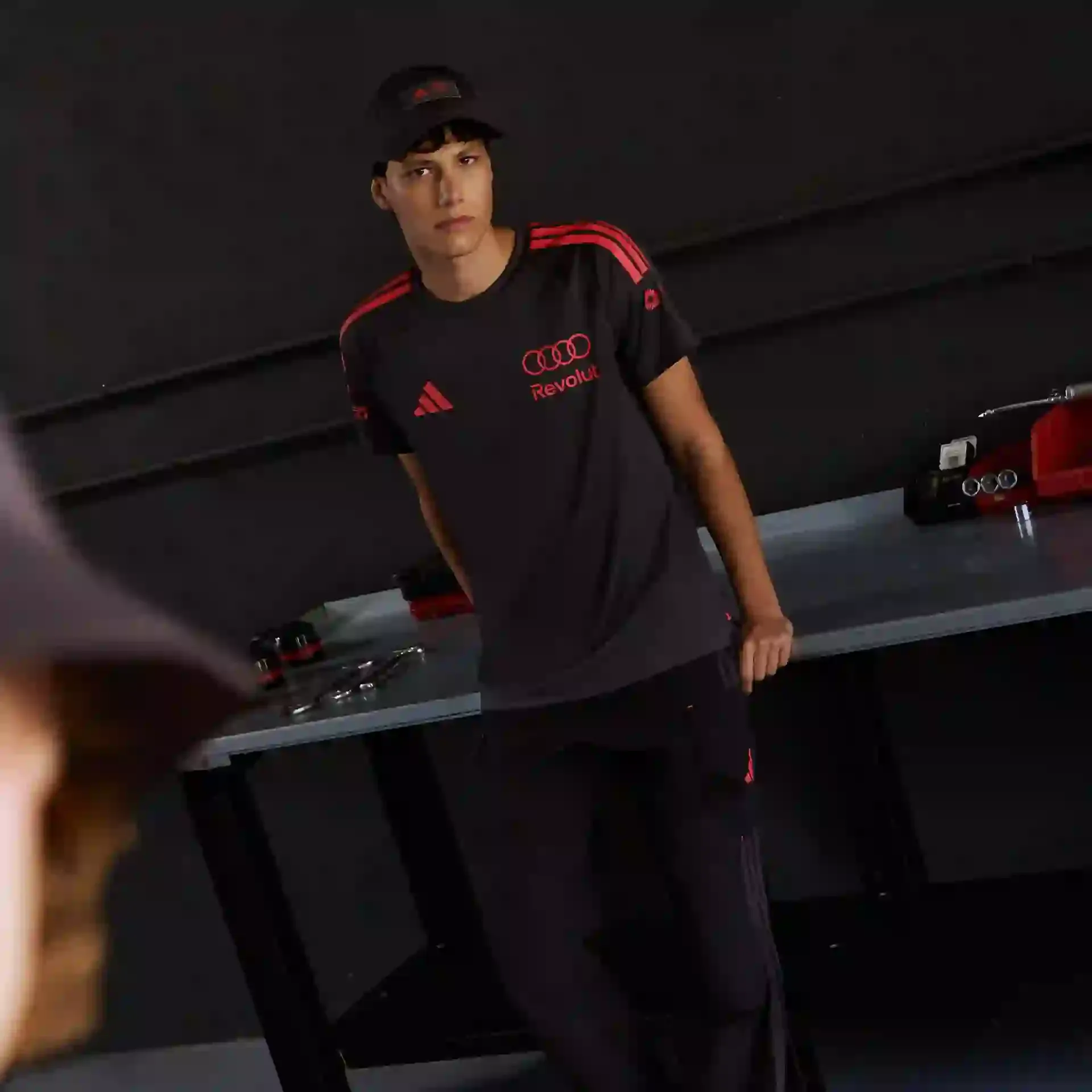 Model in dark team outfit standing, black tshirt with red adidas Logo and Audi rings Logo leaning against a mechanics work station, race track garage environment focus on athletic silhouette and clean lines