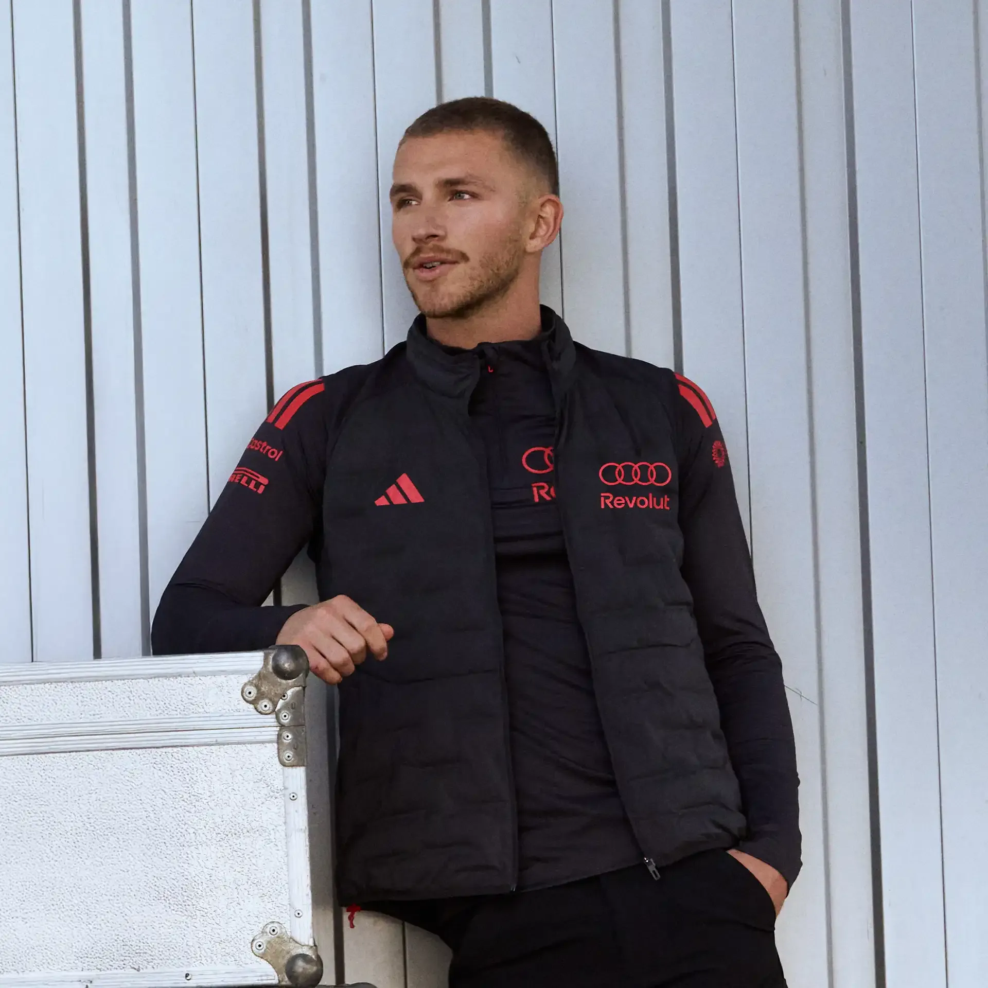 Male model leaning against wall, dark mechanic vest, with red Adidas and Audi ring Logo, inside the garage at a racing track