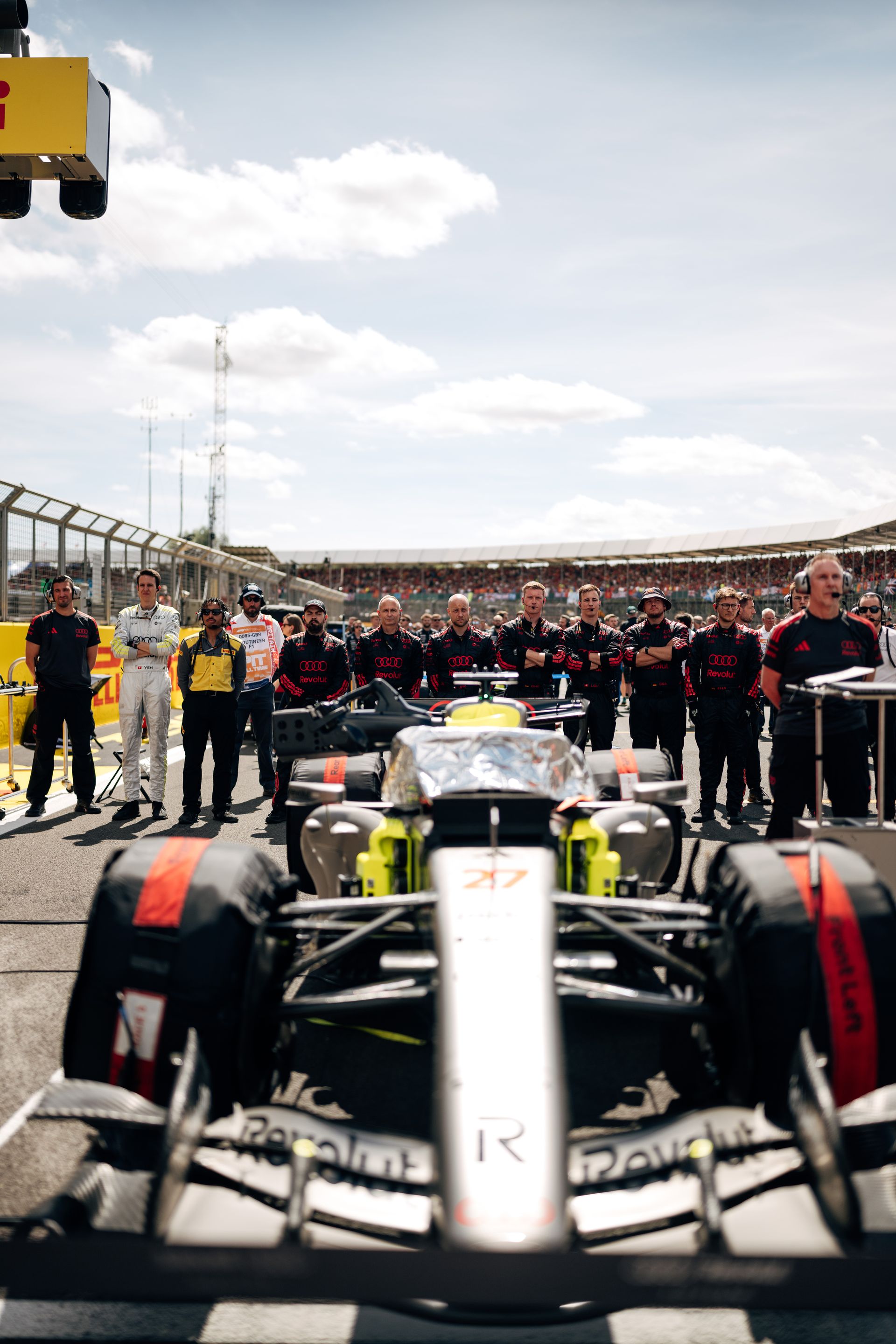 The Audi R26 is positioned on the Silverstone grid with Audi Revolut F1® Team crew lined up behind, moments before the race start.