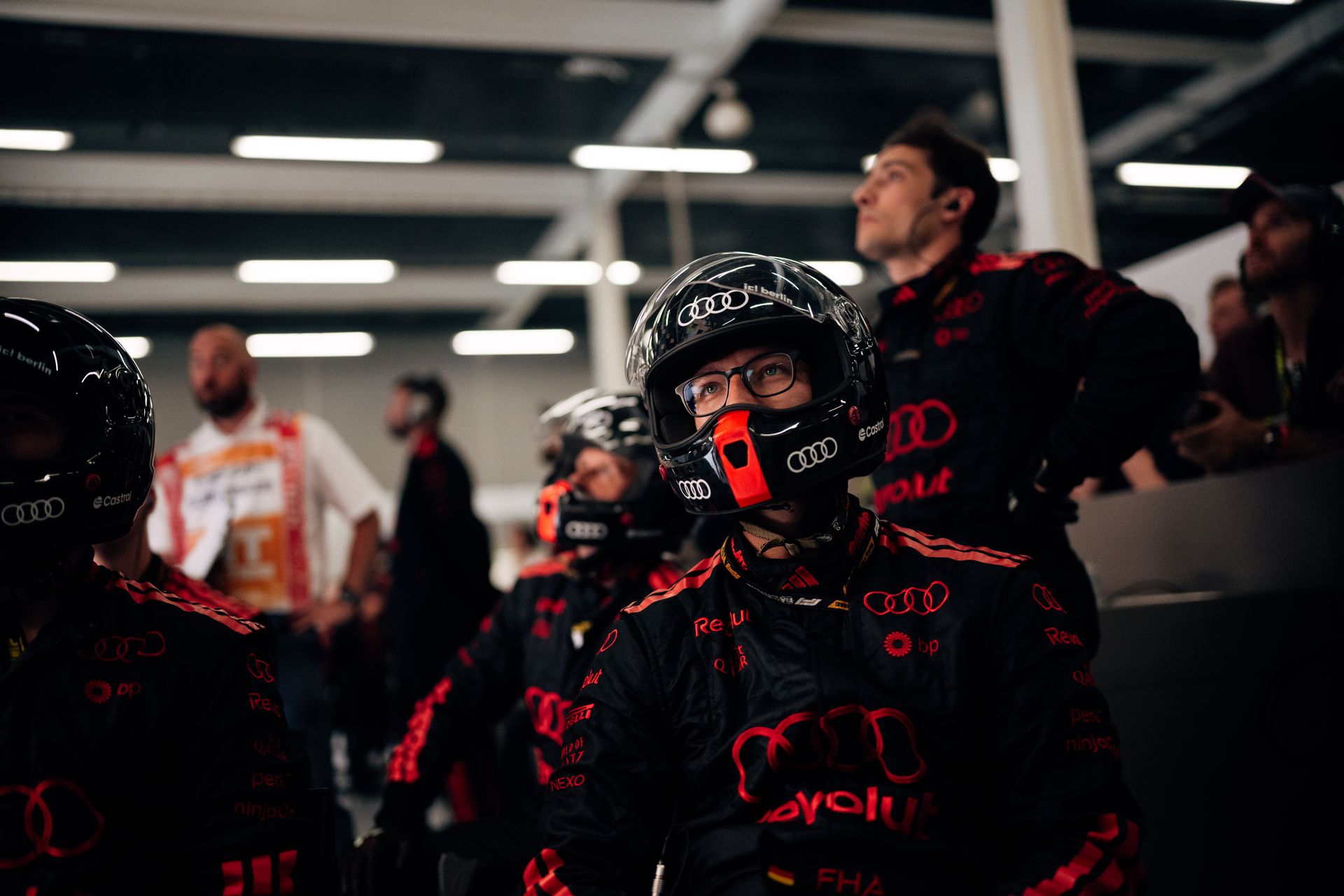 Audi Revolut F1® Team pit crew members wear helmets and watch monitors in the garage, focused on race operations.