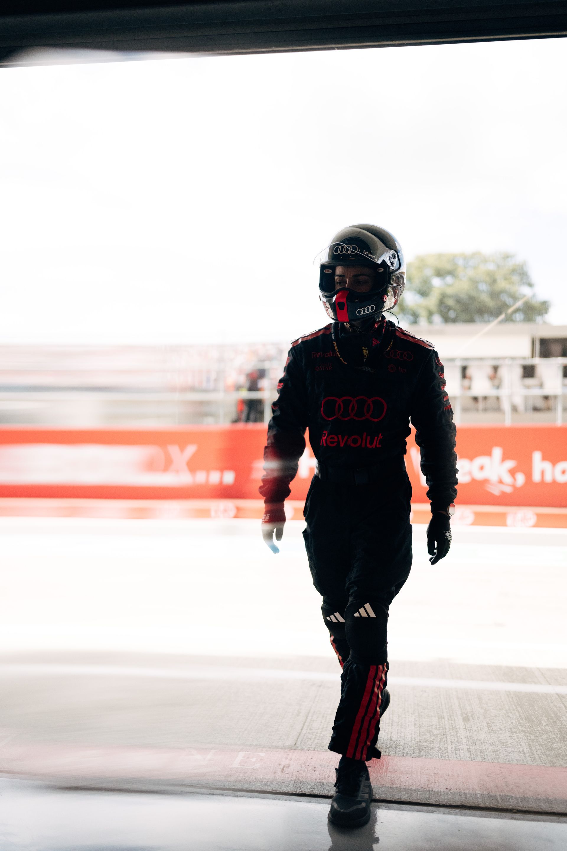 An Audi Revolut F1® Team pit crew member walks through the garage in full helmet and race gear, heading towards the pit lane.