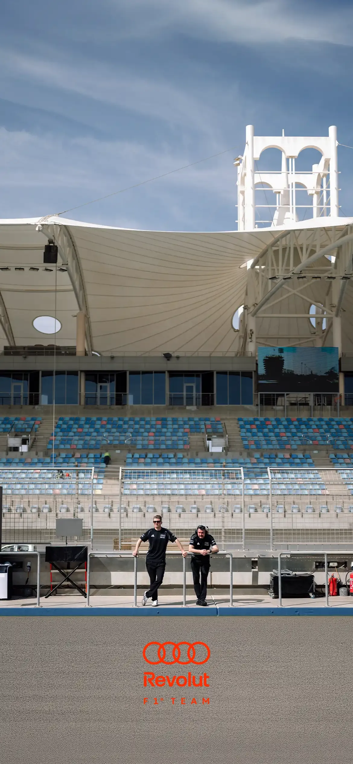 Two people of the AUDI REVOLUT F1® TEAM. Nico Hulkenberg left and James Key crewmember on the right at Testweek 01 from Bahrain, stand at the front railing of a mostly empty race track, with blue and gray seats. Above them is a large white tensile canopy roof supported by structural beams, with a tall white tower rising in the background. A large video screen is mounted on the right side, and the sky is lightly clouded