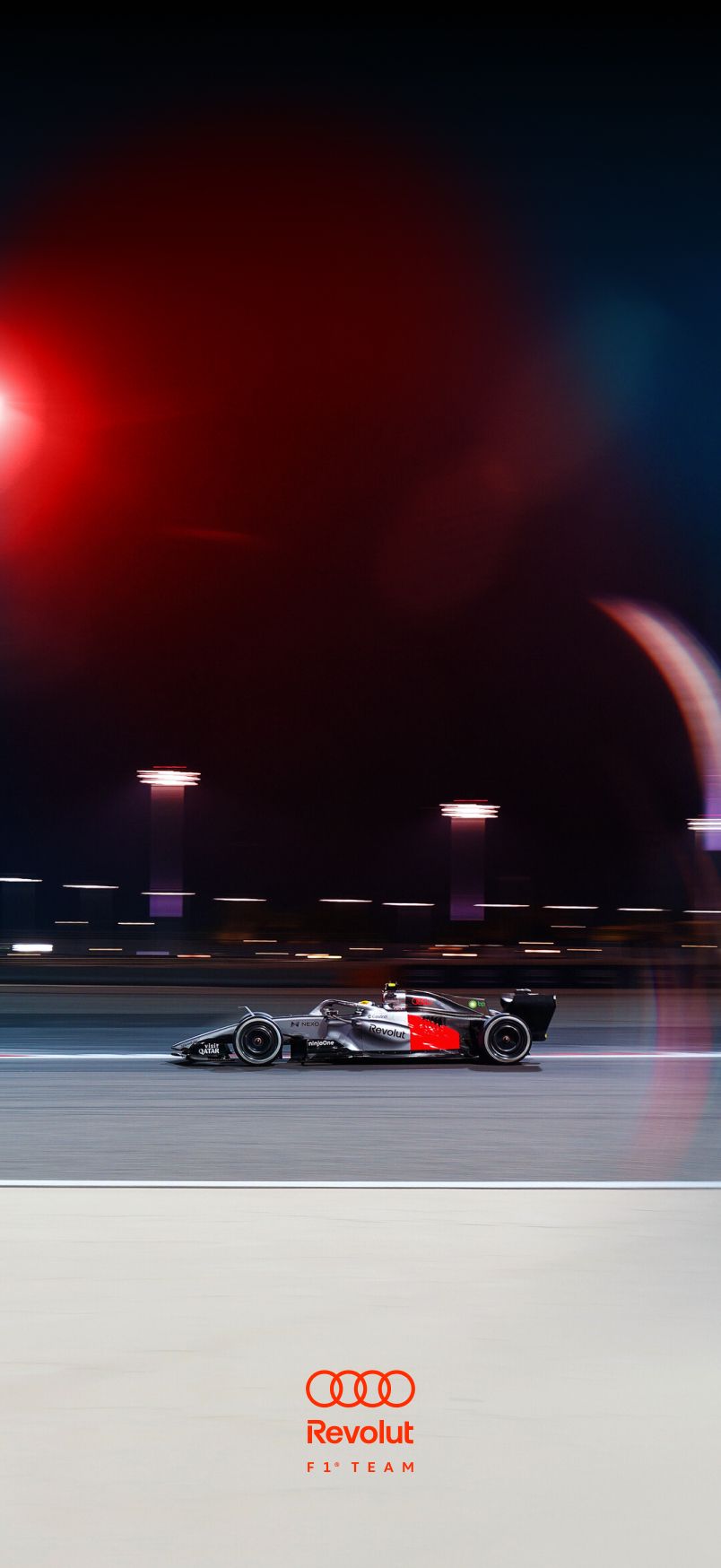 Sideview of Audi Revolut F1® Team car race car on a racetrack at night with motion blur on a floodlit circuit; red and blue light reflections and track lamps create a strong sense of speed and movement.