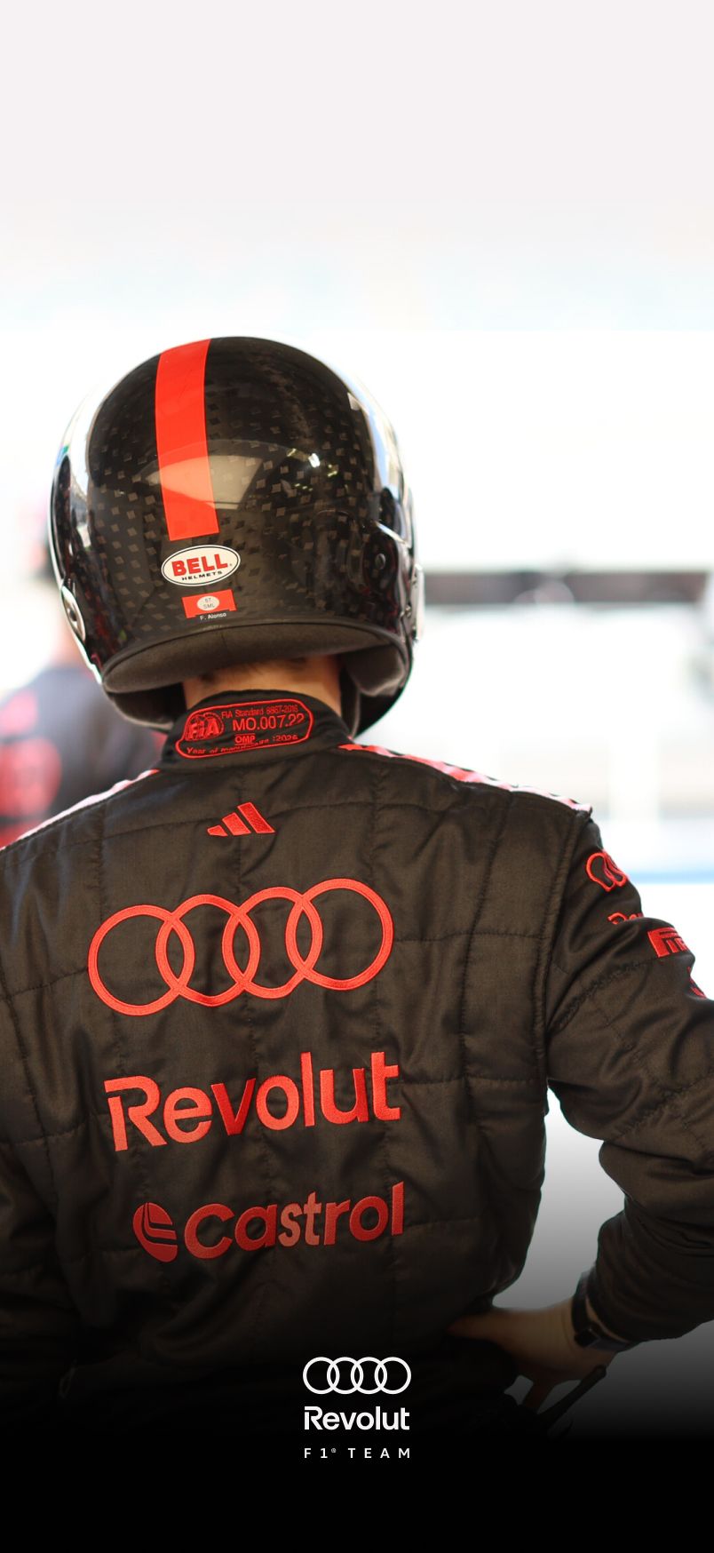 Rear view of a racing driver of the Audi Revolut F1® Team wearing a black Audi motorsport suit and helmet, featuring red Audi rings and sponsor logos including Revolut and Castrol, standing in a garage environment.