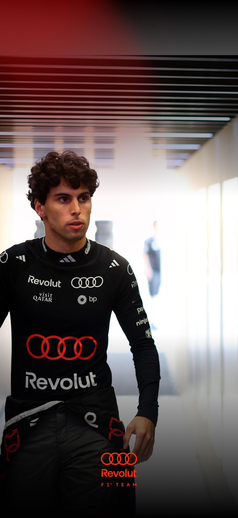 Portrait of a young male racing driver Gabriel Borotelo of the Audi Revolut F1® Team with curly hair wearing a black Audi-branded racing suit with sponsor logos, standing indoors and looking focused off-camera.