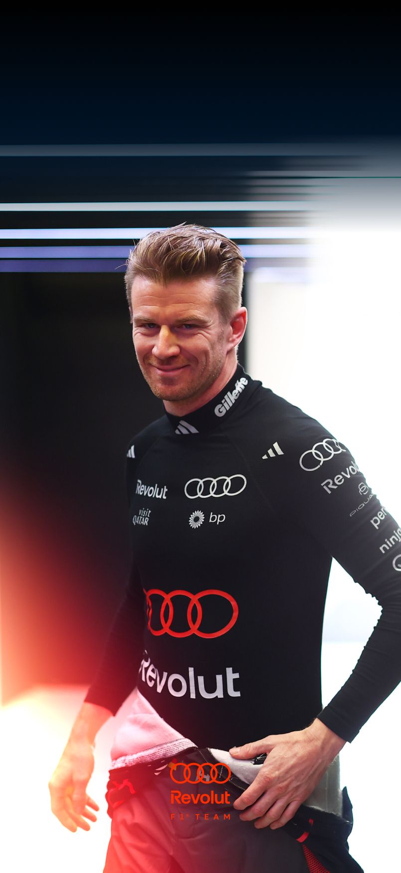 Portrait of a male racing driver Nico Hulkenberg of the Audi Revolut F1® Team blond short hair, wearing a black Audi-branded racing suit with sponsor logos, standing indoors and looking focused off-camera.