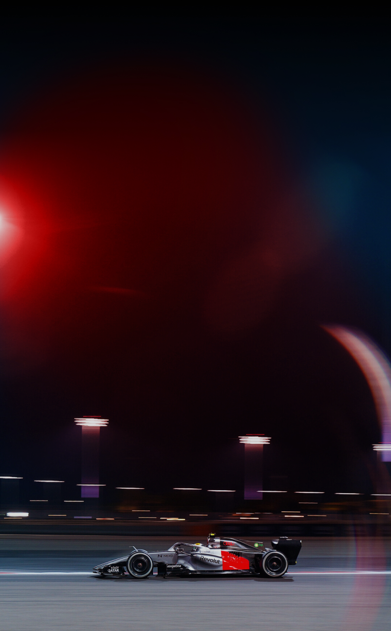 Sideview of Audi Revolut F1® Team car race car on a racetrack at night with motion blur on a floodlit circuit; red and blue light reflections and track lamps create a strong sense of speed and movement.