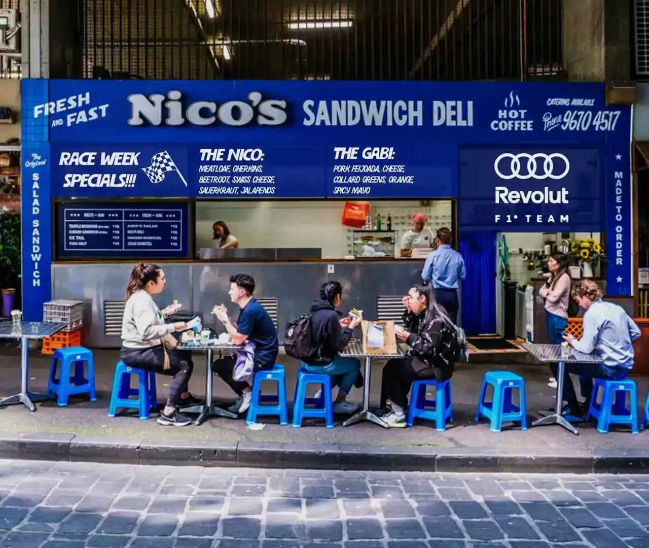 An image of Nicos Deli front view, a lot of people sitting and eating at the tables, urban style Deli