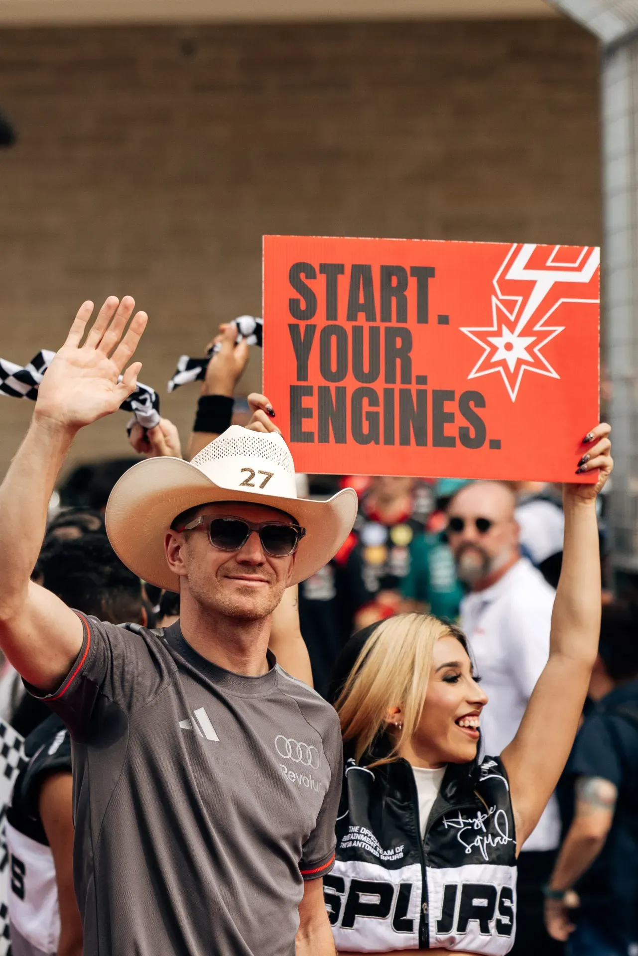 Man in a cowboy hat with number 27 waves beside a woman holding a Start Your Engines sign in Austin