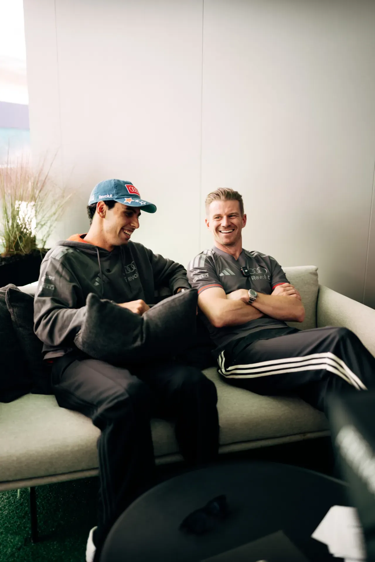 Gabriel Bortoleto and Nico Hulkenberg of the Audi Revolut F1® Team sit on a sofa, one in a blue cap holding a cushion, one with arms crossed, in Austin.