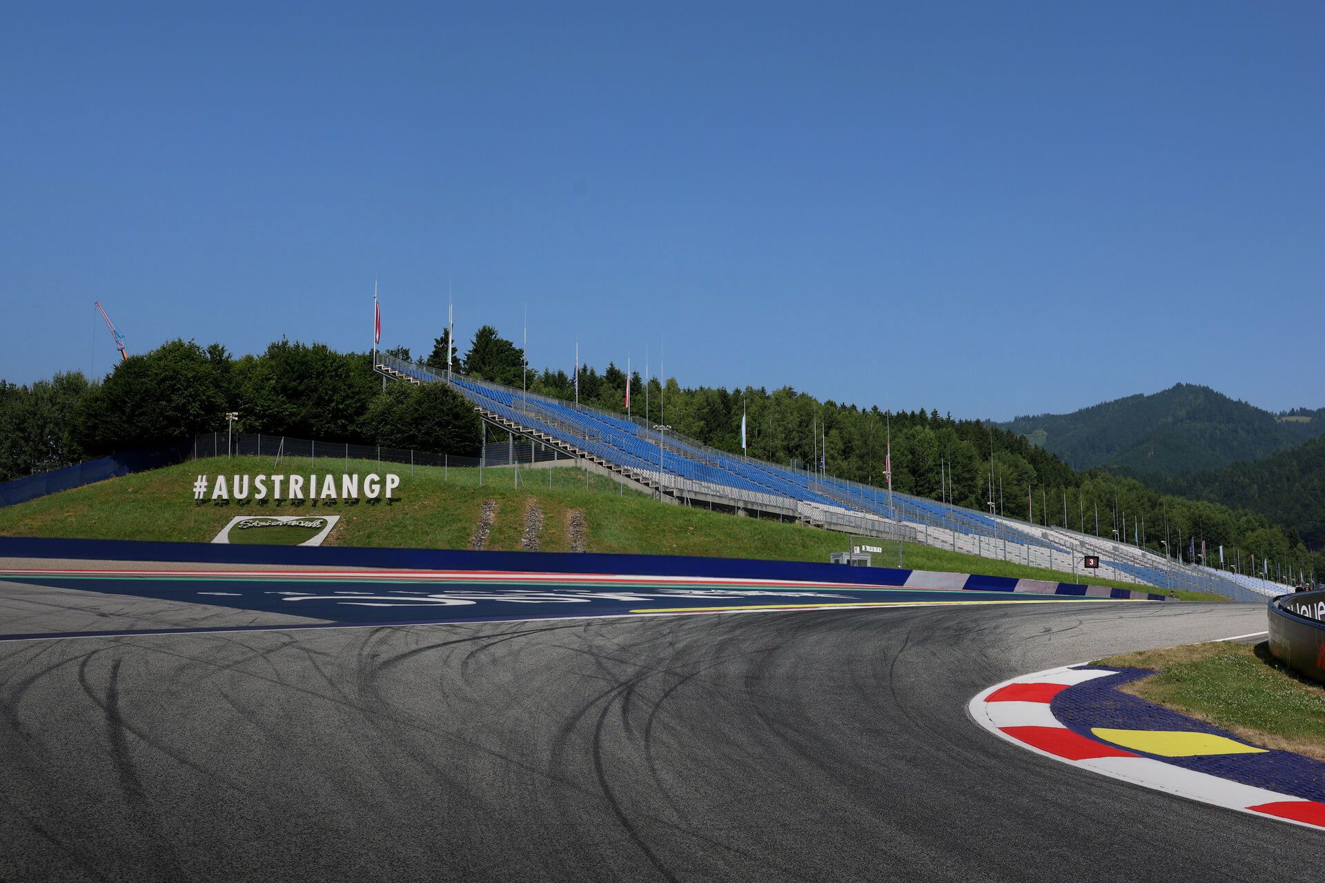 A sweeping corner at the Red Bull Ring in Spielberg, Austria, with tire marks on the asphalt, red-white-blue kerbs, empty grandstands, and forested hills under a clear blue sky.