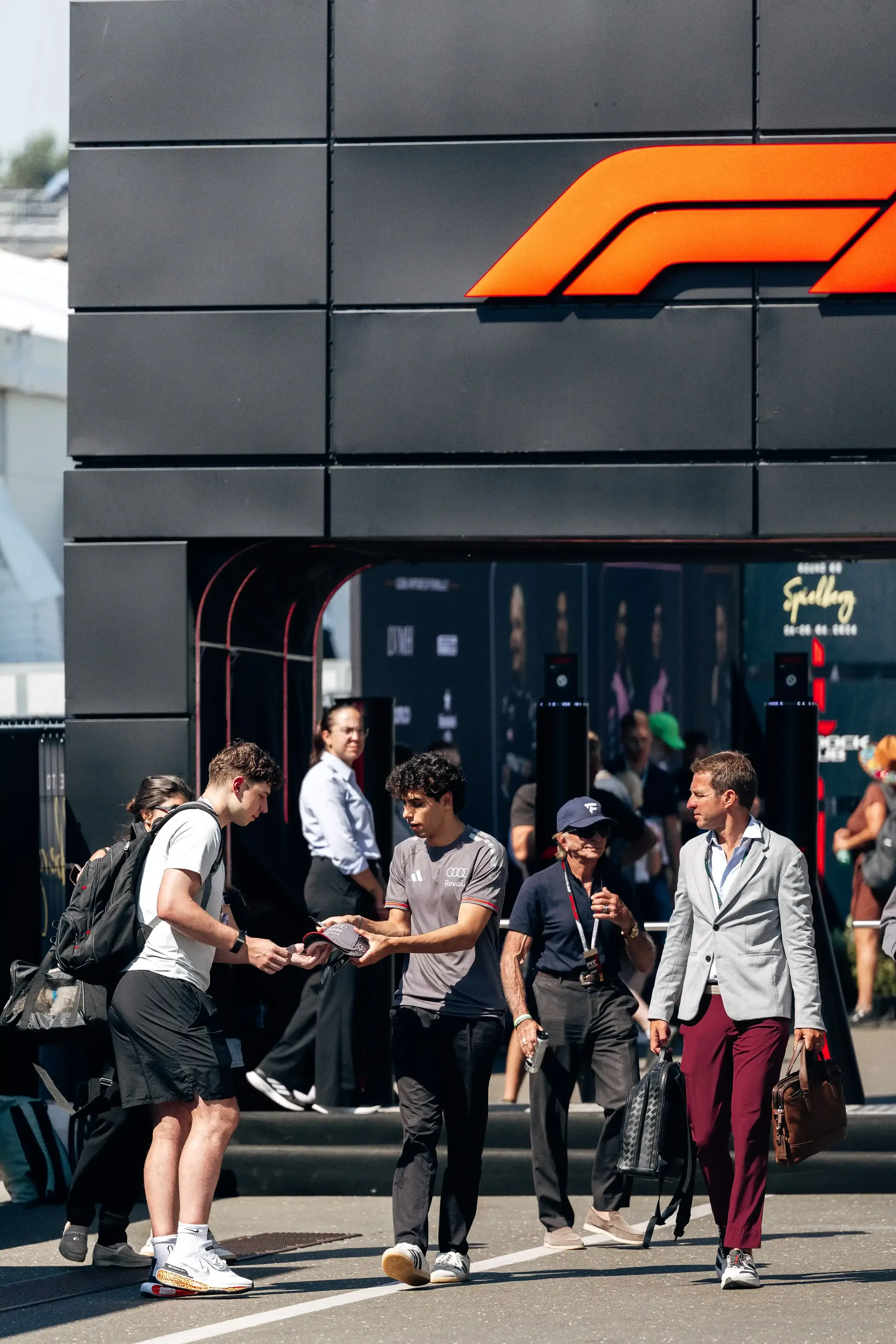 Gabriel Bortoleto driver at the Audi Revolut F1® Team walk past the entrance of a paddock building featuring a large orange team logo.