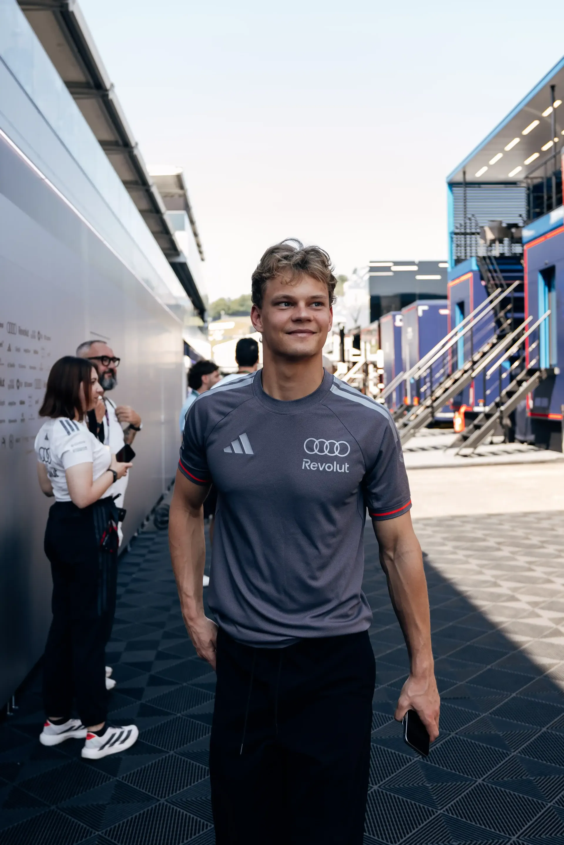 Paul Aron driver at the Audi Revolut F1® Team smiles and walks along the paddock lane between hospitality and garage structures; this image is currently selected and outlined.