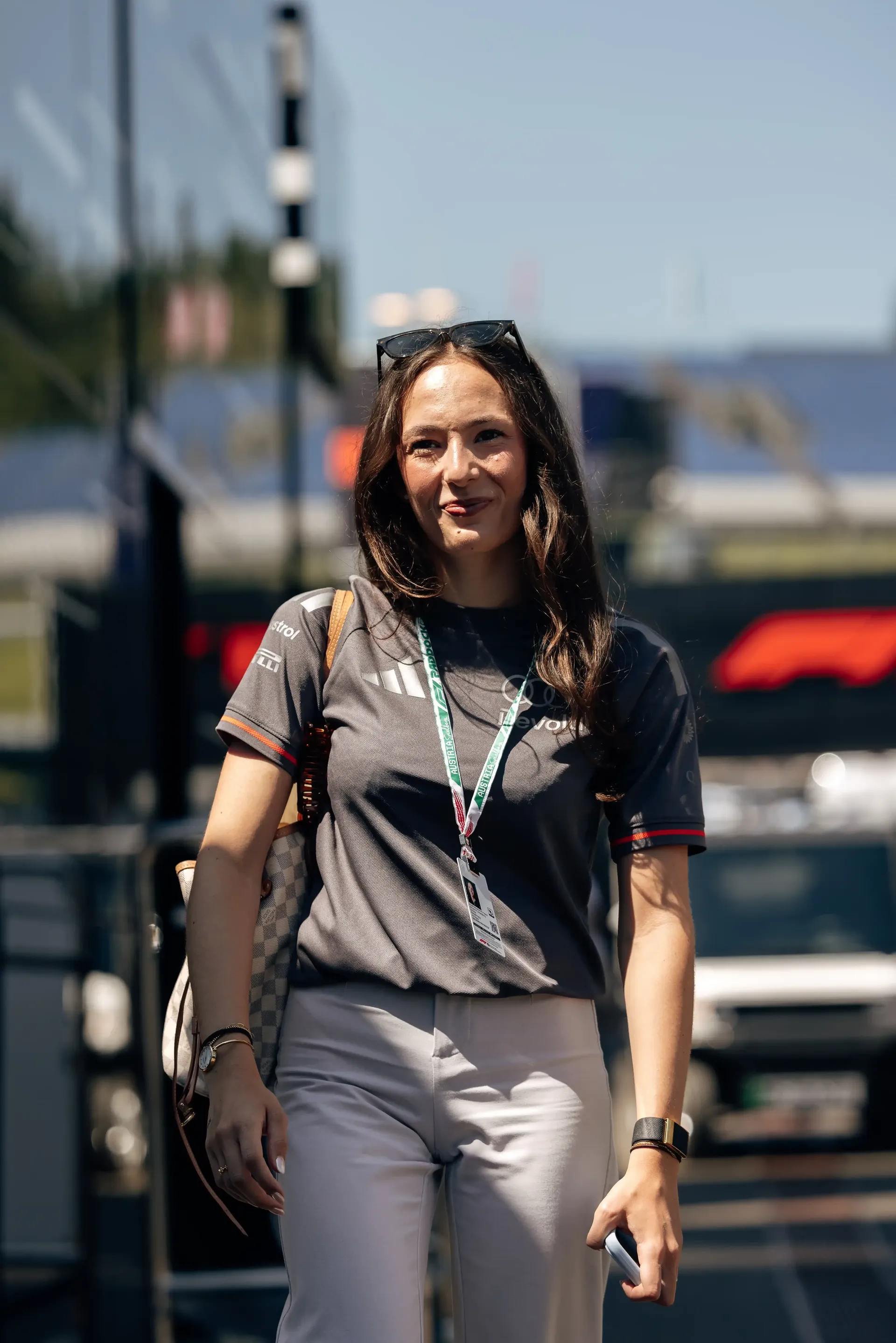 Emma Felbermayr driver at the Audi Revolut F1® Team Academy wearing a lanyard walks through the paddock, with support vehicles and circuit infrastructure visible behind her.