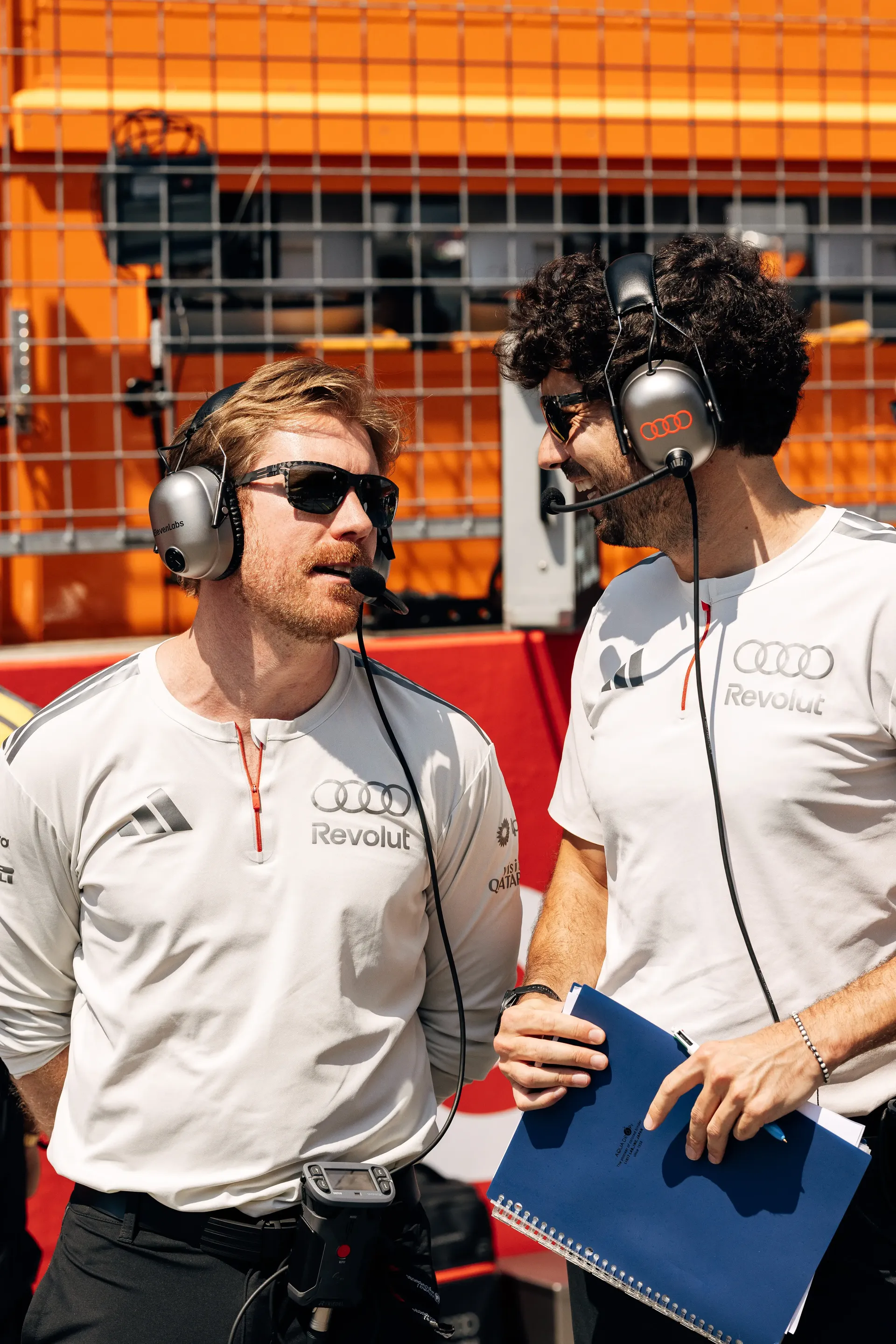 Two Audi Revolut F1® Team engineers wearing white team shirts and headsets discuss race operations in the pit lane.