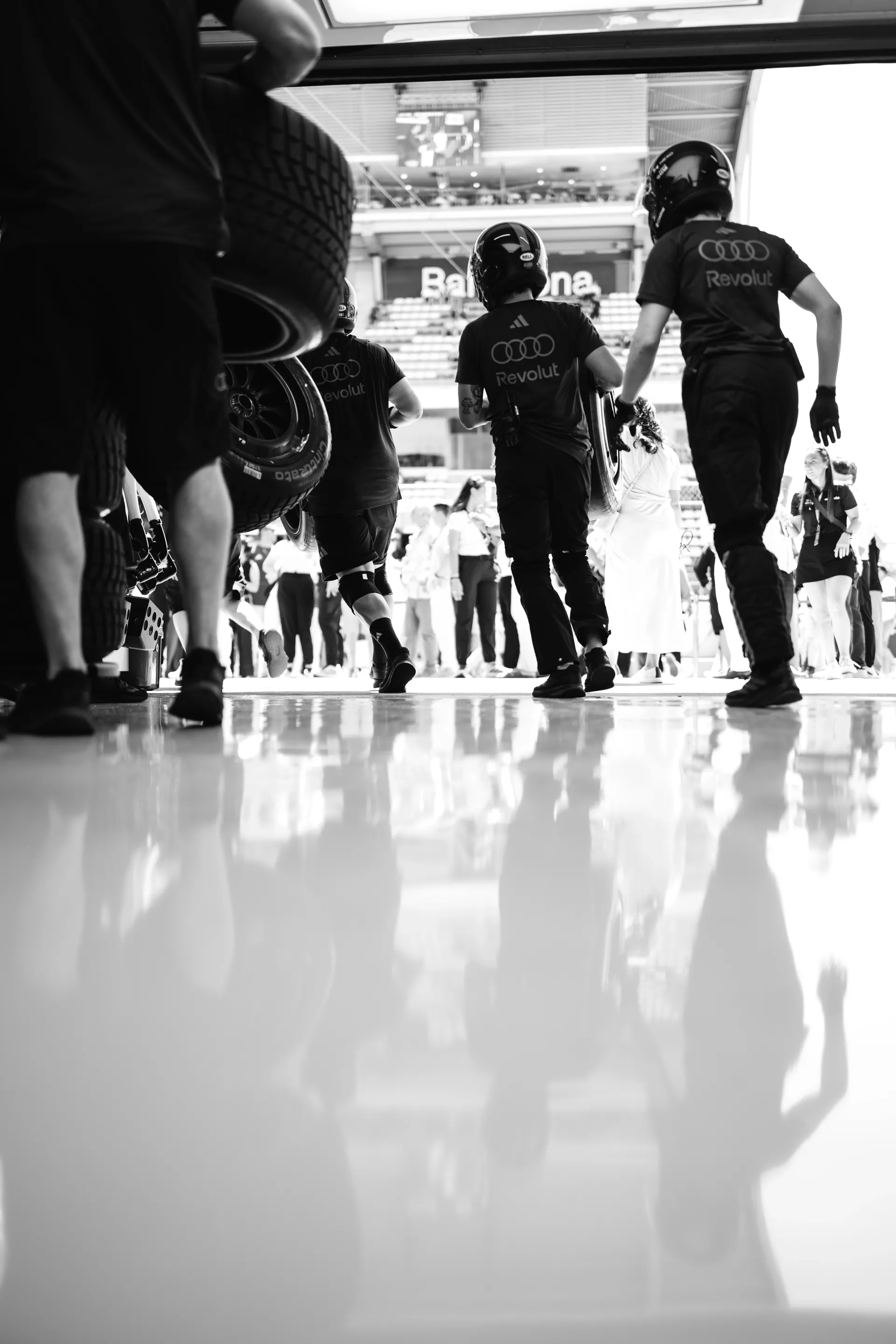 Audi Revolut F1® Team pit crew members run out of the garage carrying tires during a pit stop rehearsal, shot from a low angle with reflections on the floor