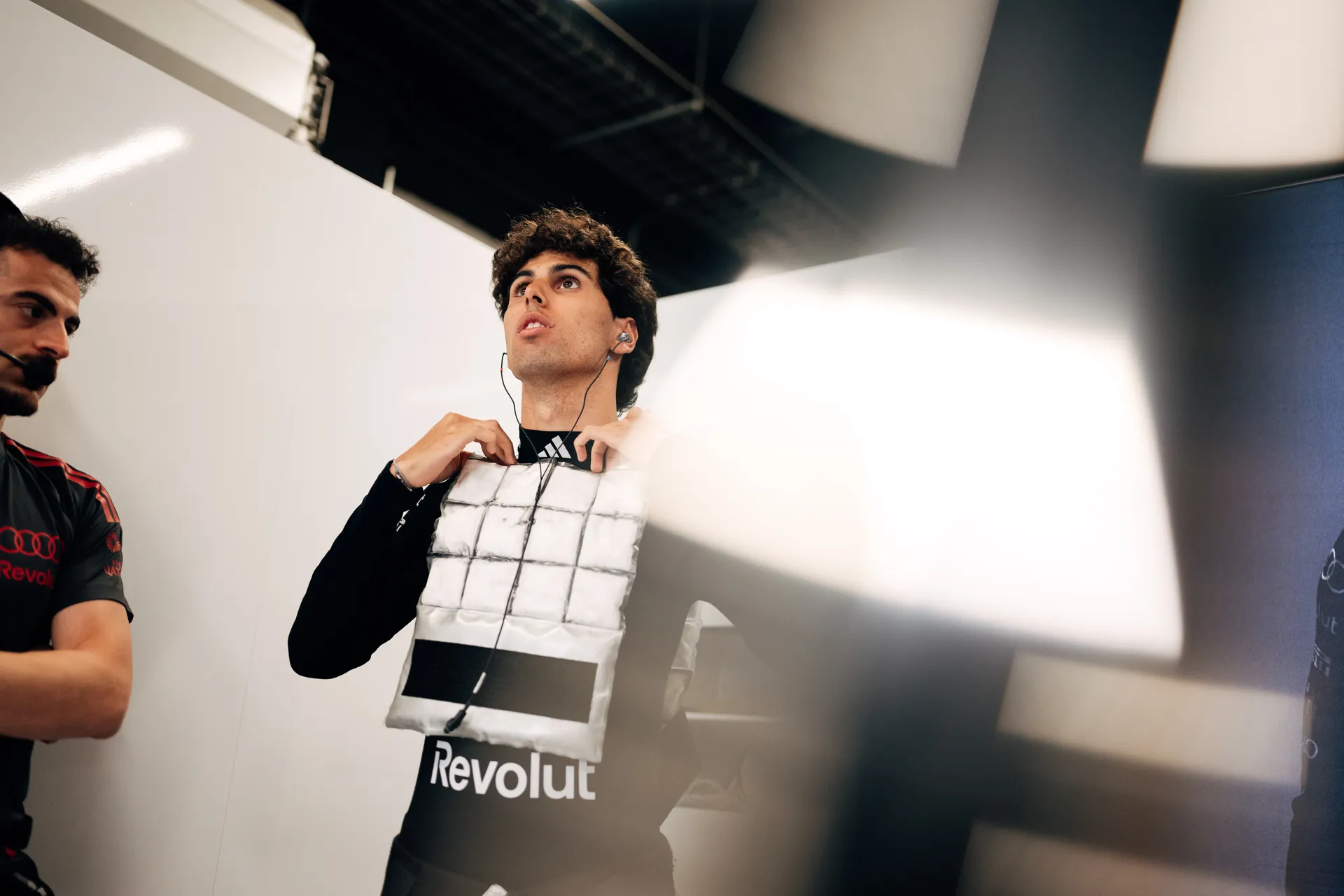 Gabriel Bortoleto adjusts his protective vest over his race suit with in-ear monitors, preparing to get into the car inside the garage
