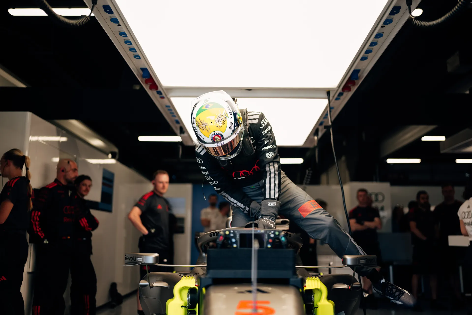 Gabriel Bortoleto climbs into car number 5 inside the Audi Revolut F1® Team garage as crew members observe