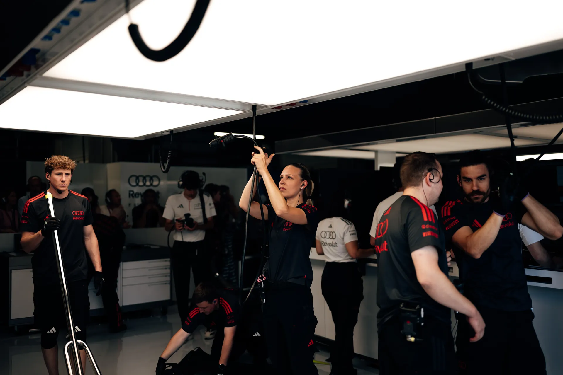 Audi Revolut F1® Team crew members work inside the garage, adjusting overhead equipment and preparing for the next run while Gabriel Bortoleto stands to the side