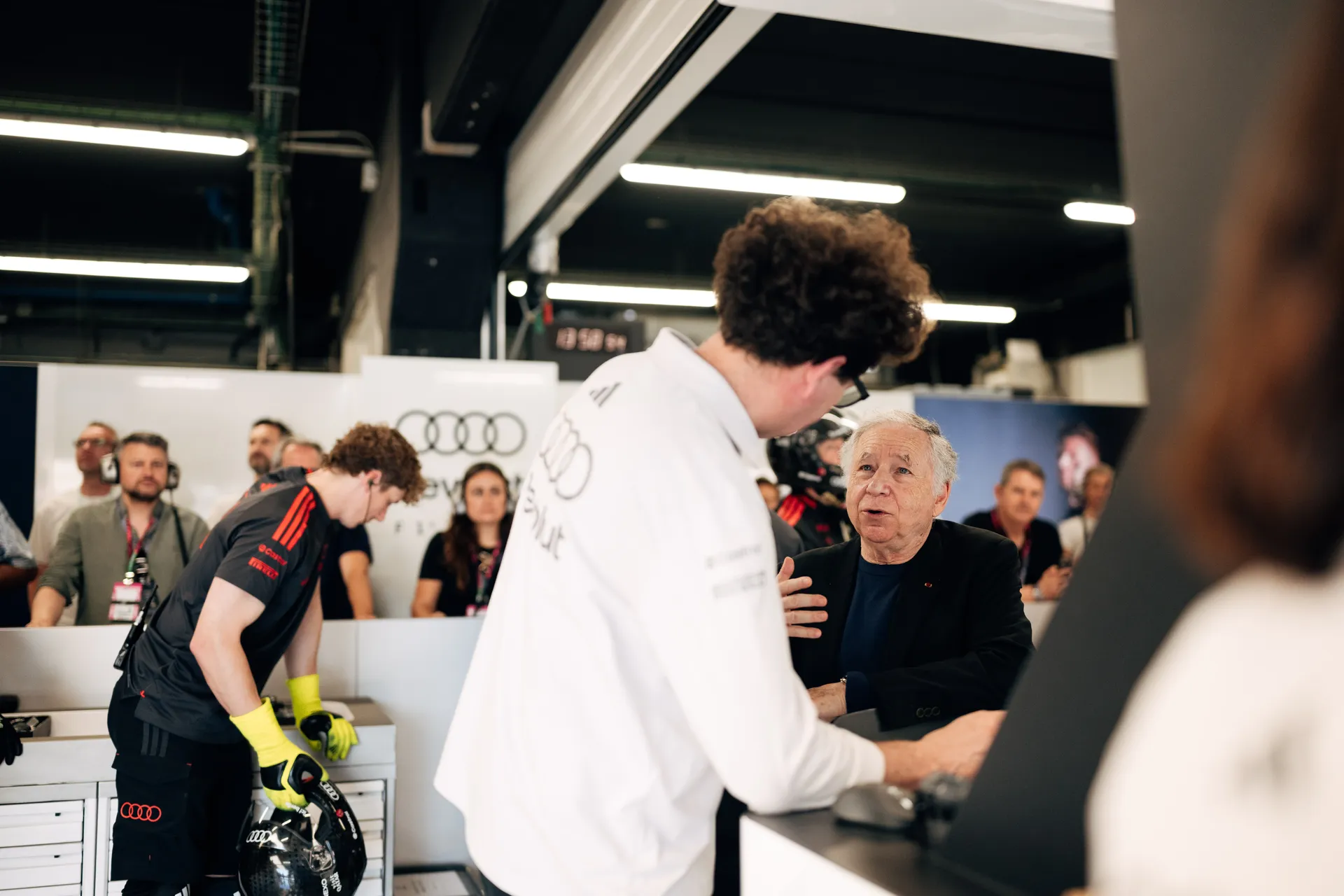 Audi Revolut F1® Team garage scene with team members in discussion at the pit wall while a mechanic prepares equipment in the background.