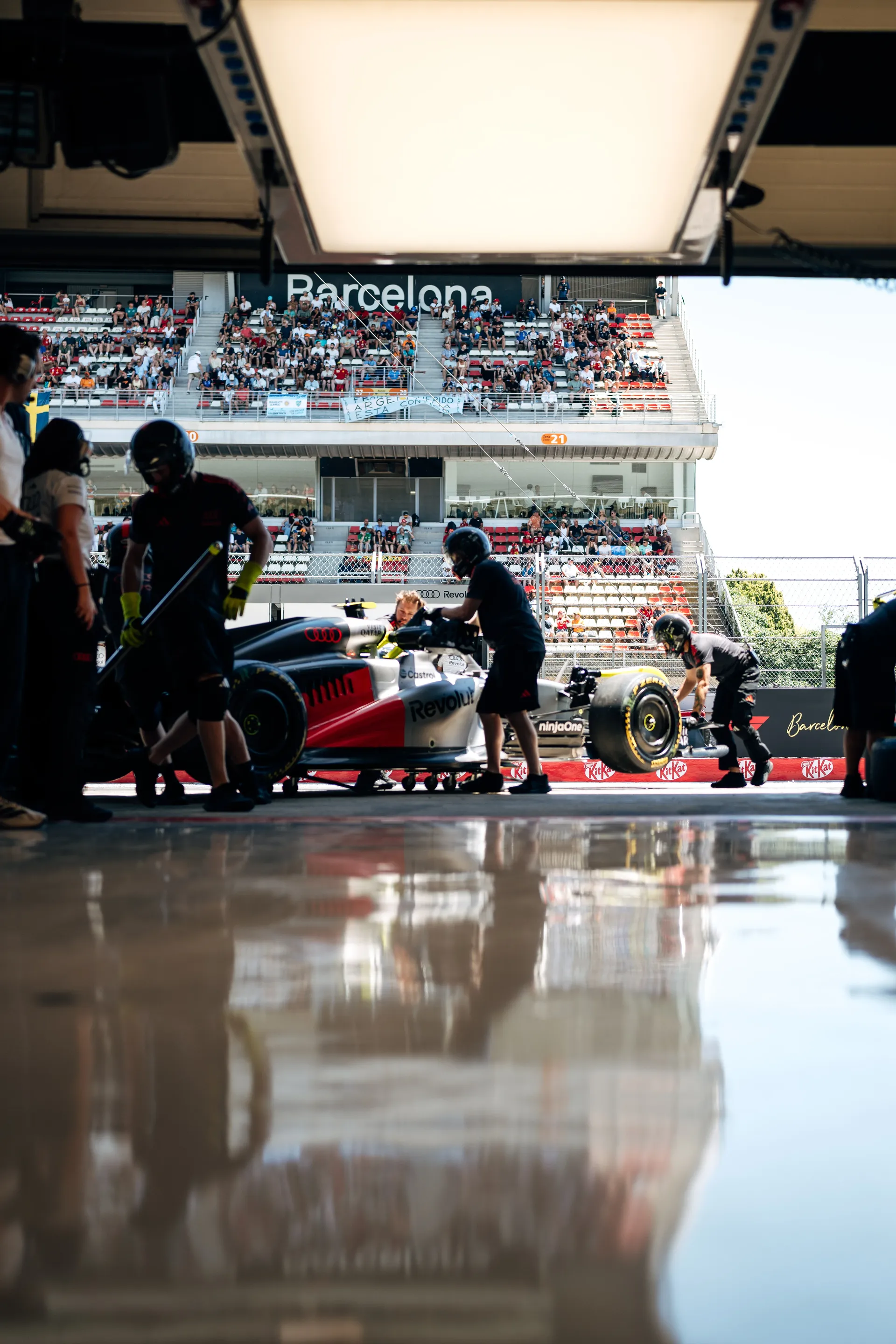 Audi Revolut F1® Team car being pushed out of the garage during FP1 in Barcelona, with the grandstand and spectators visible opposite the pit lane.