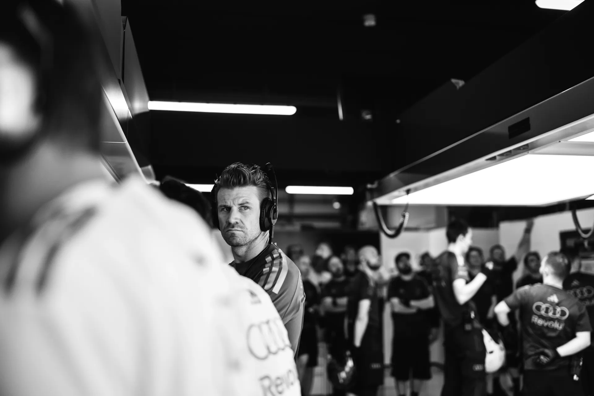 Black and white portrait of an Audi Revolut F1® Team engineer wearing a headset, focused inside the busy garage environment.