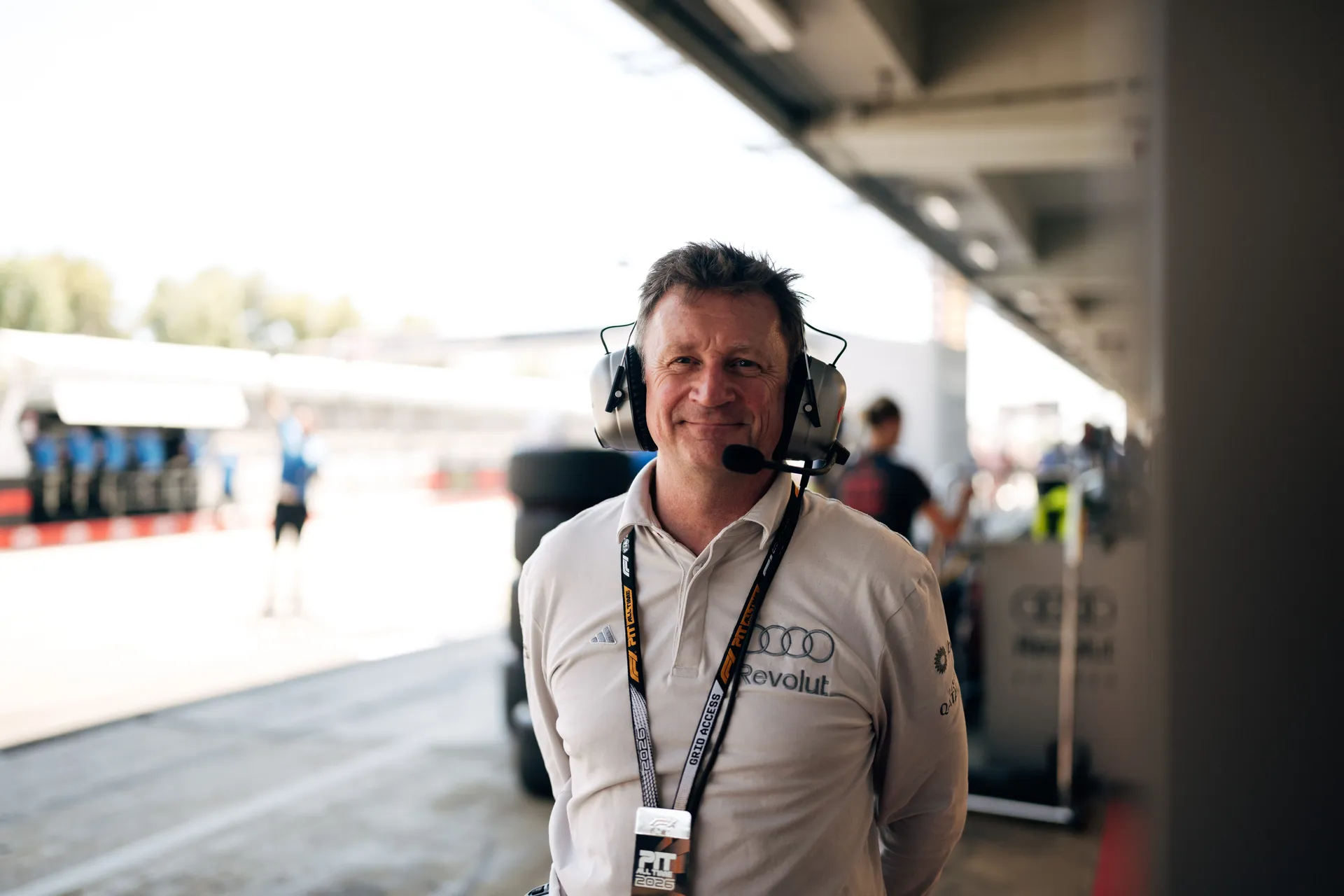 Portrait of Allan McNish Audi Revolut F1® Team engineer wearing a headset and smiling in the pit lane environment.