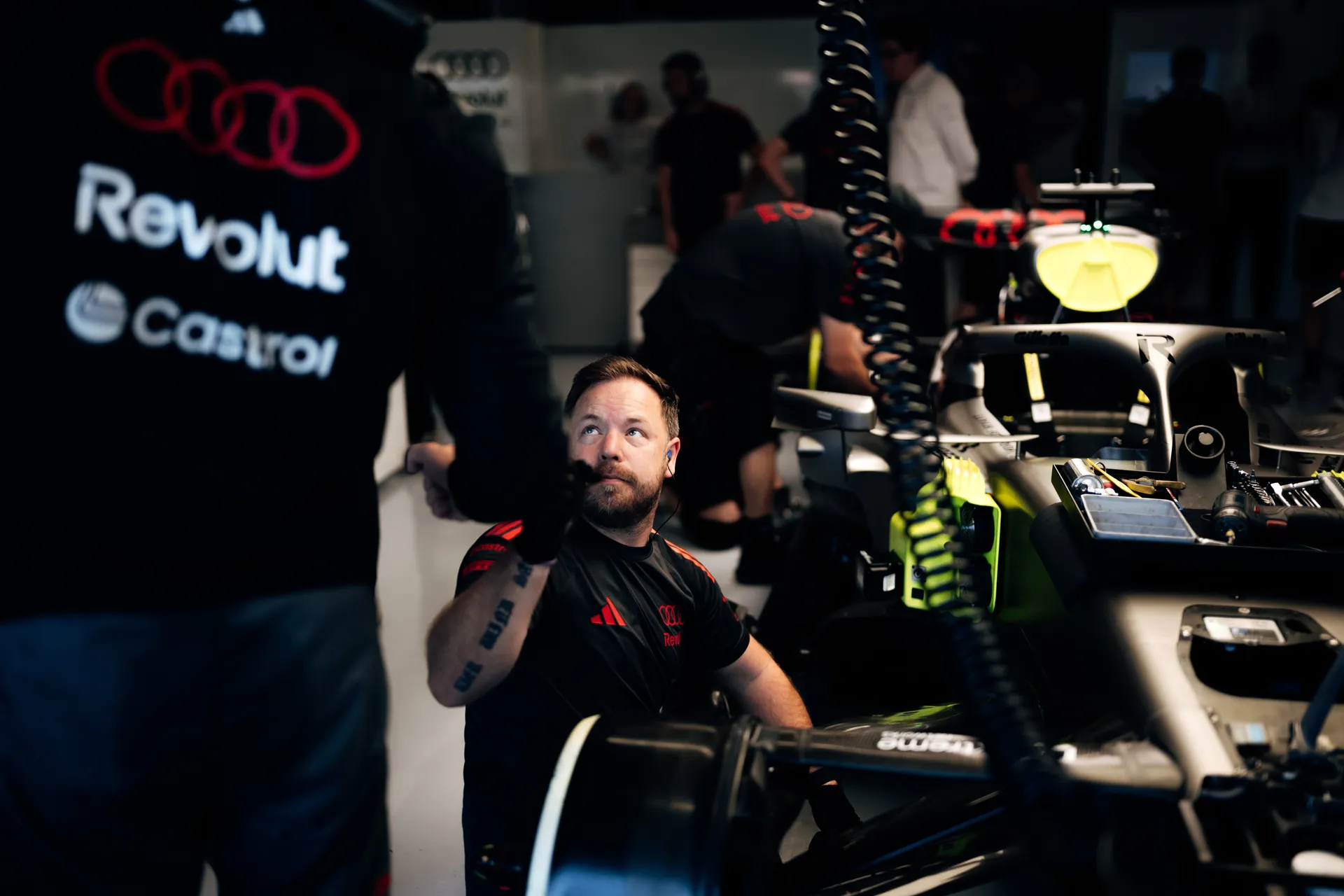 Audi Revolut F1® Team mechanic signaling upward while working beside the car, with front suspension and tooling visible.