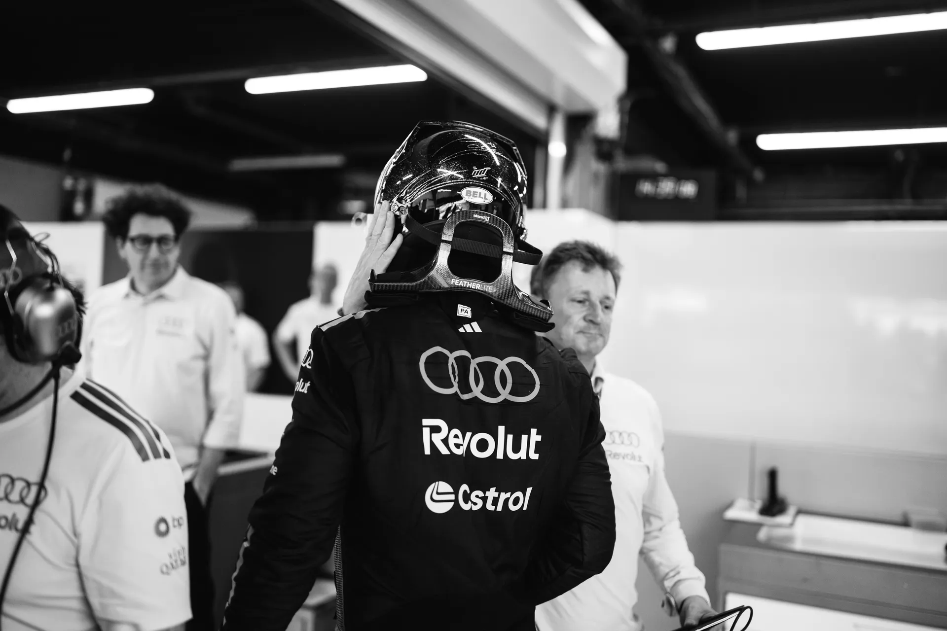 Paul Aron seen from behind adjusting his helmet while team members stand nearby in the garage.
