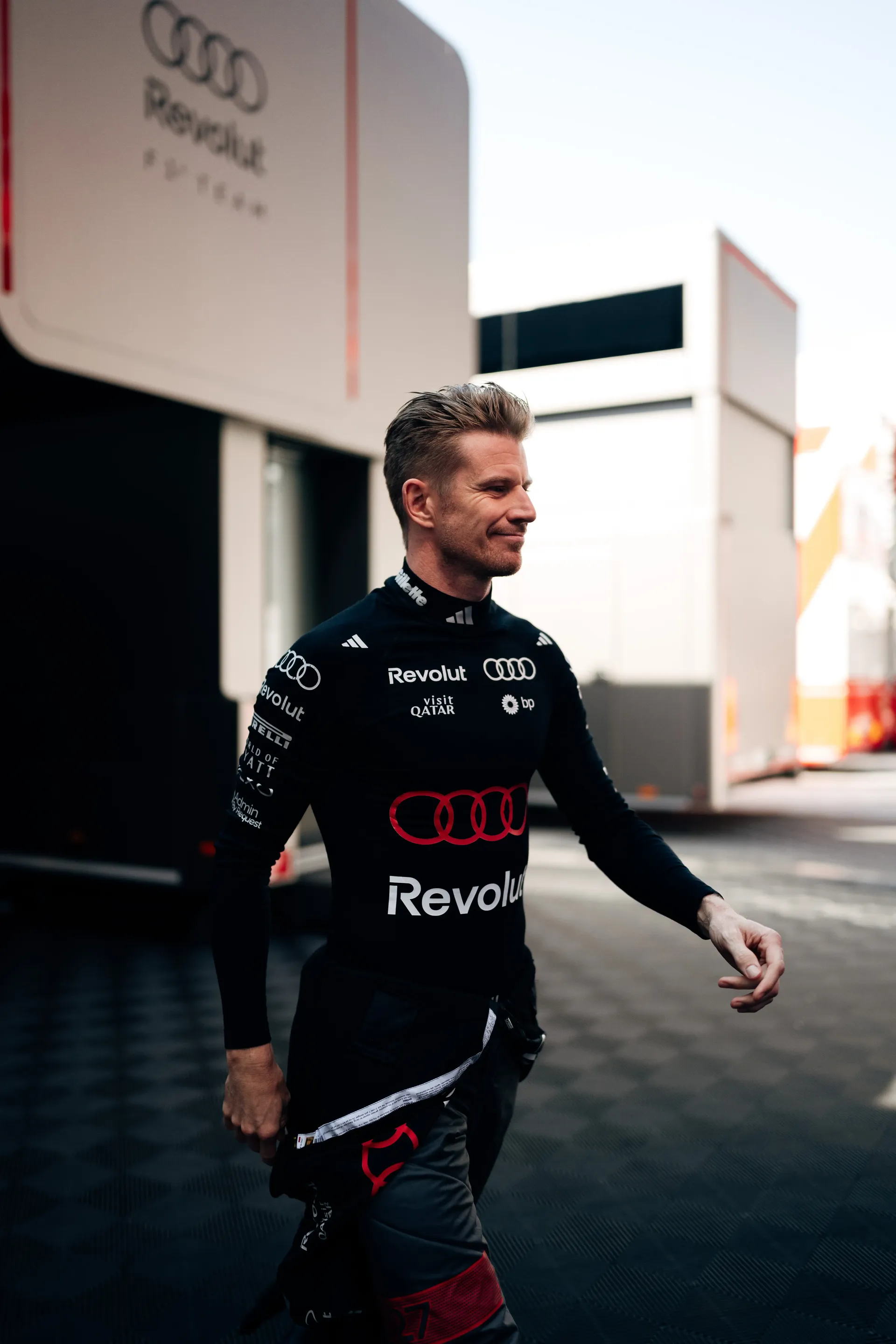 Nico Hulkenberg walking through the paddock in Audi Revolut F1® Team race suit, passing team hospitality units in the background.