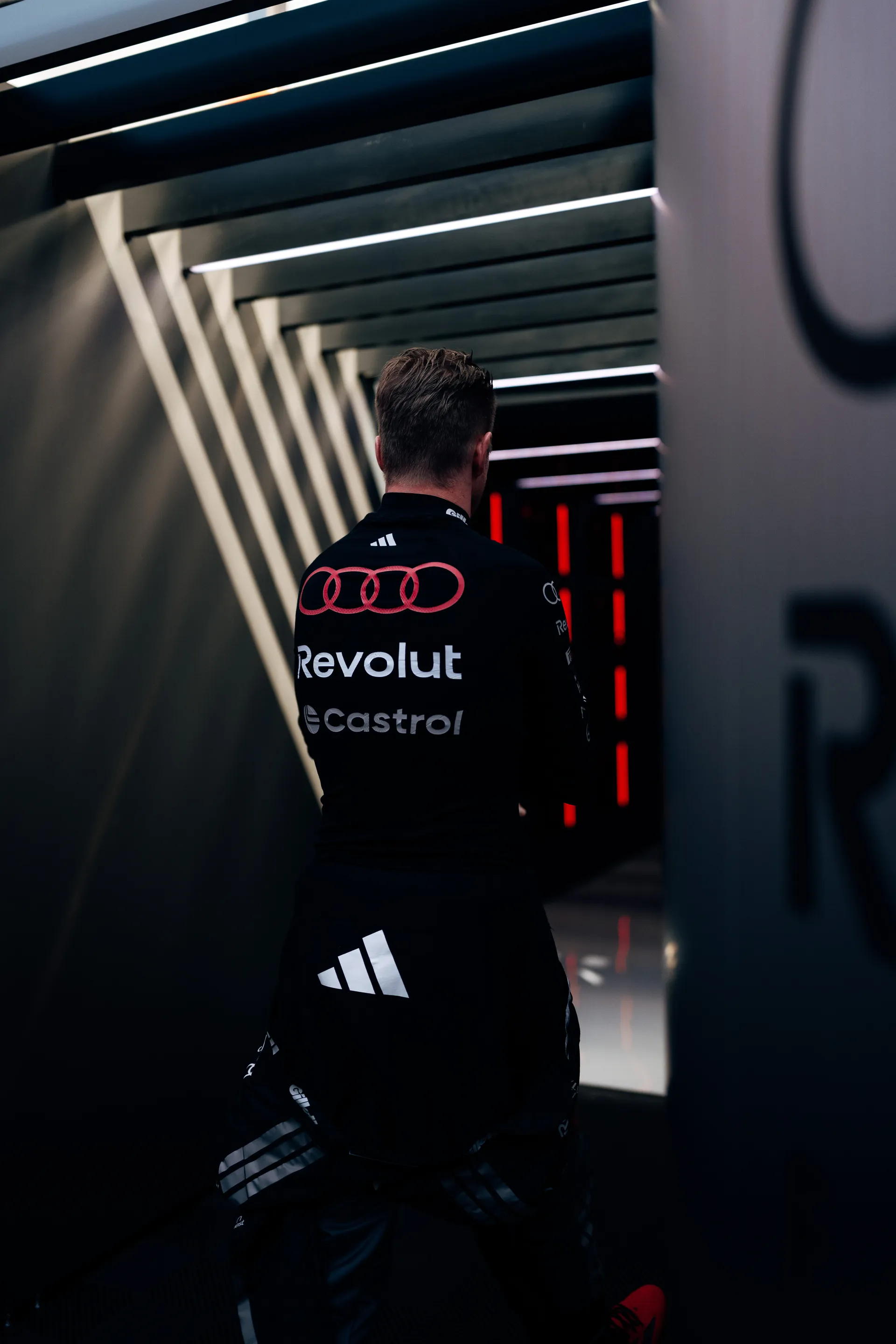 Rear view of Nico Hulkenberg walking through a lit corridor inside the Audi Revolut F1® Team garage, with geometric light structures framing the walkway.