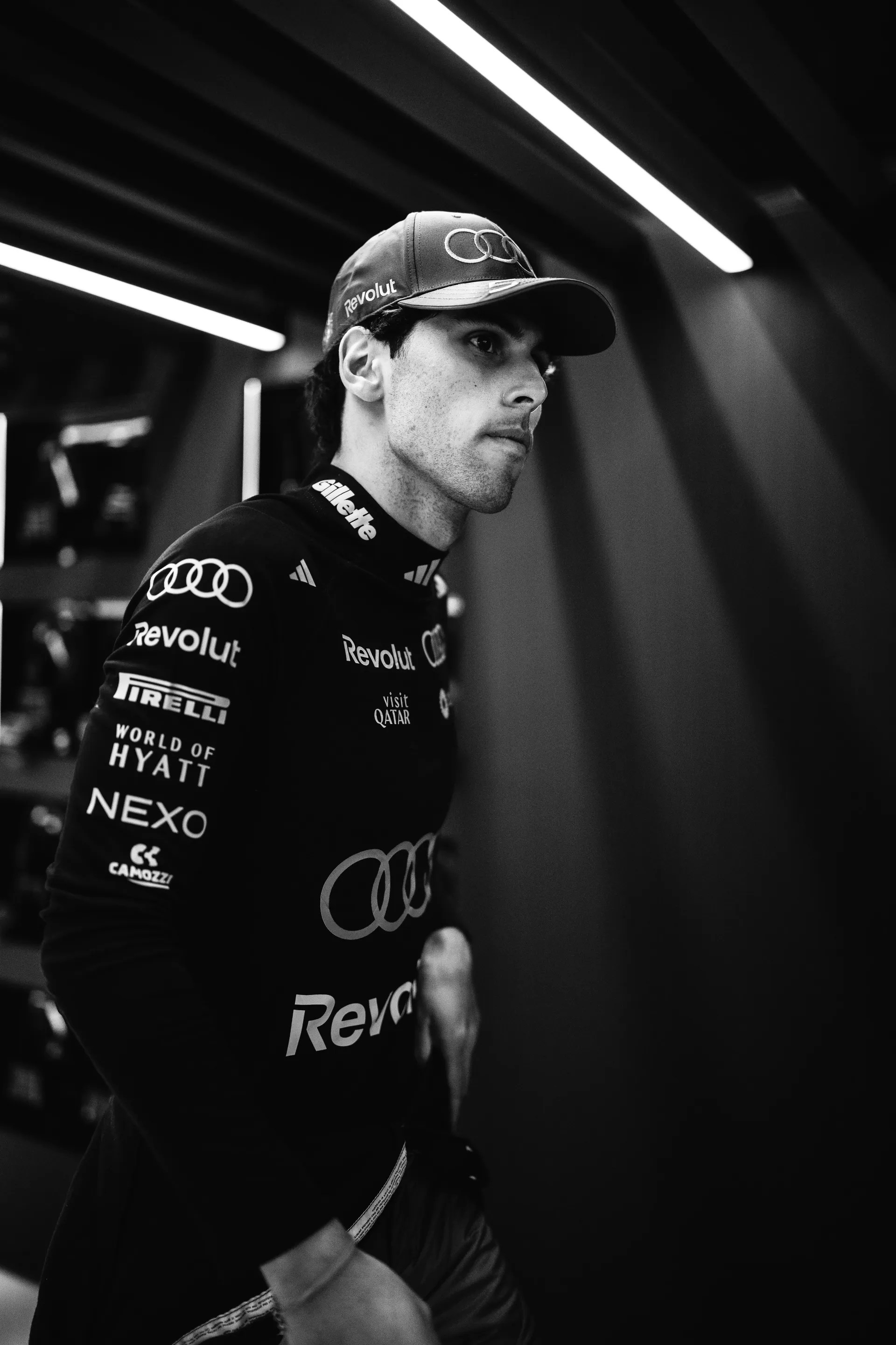Gabriel Bortoleto in black and white portrait, wearing Audi Revolut F1® Team cap and race suit, walking through the garage with focused expression.