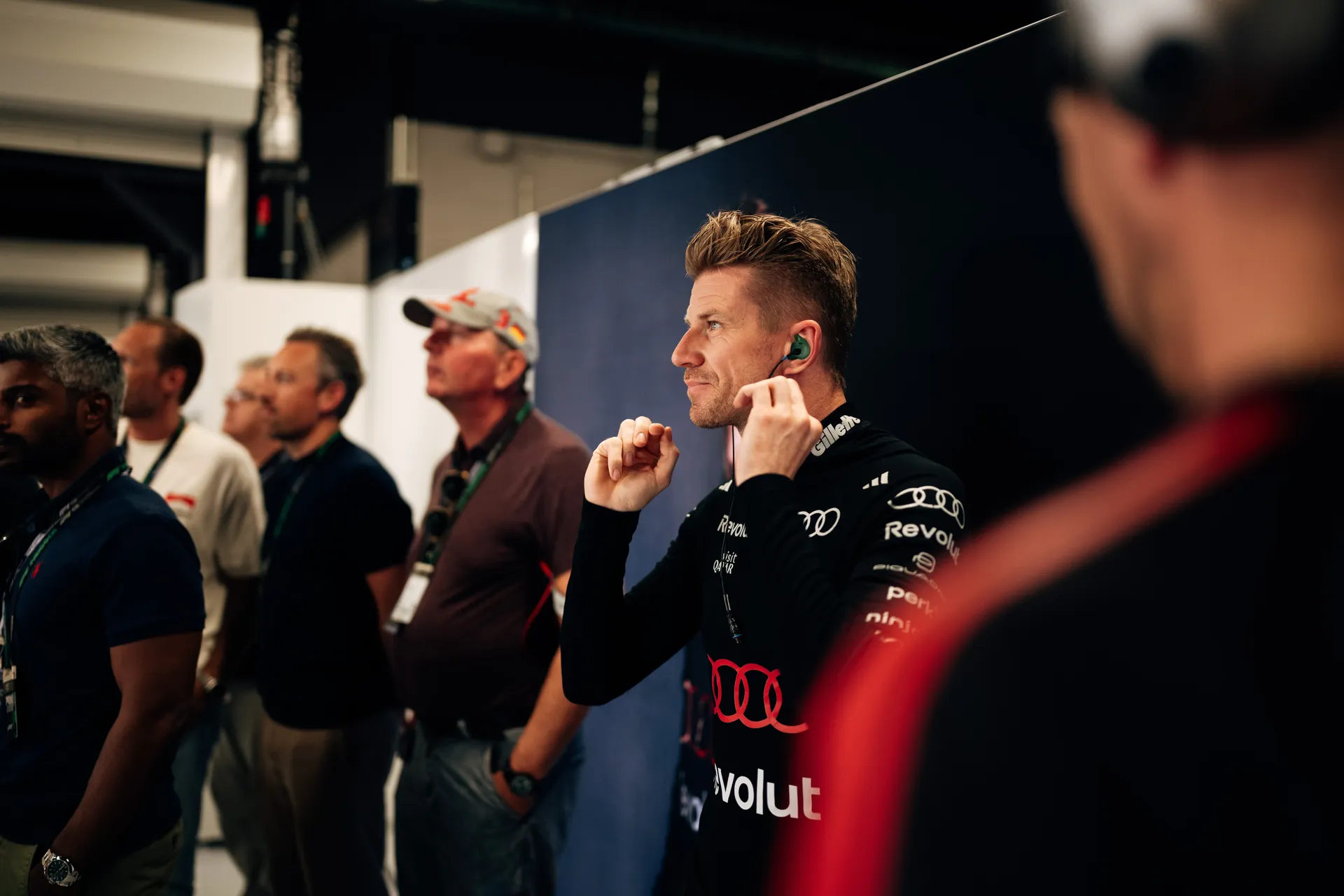Nico Hulkenberg adjusting his earpiece while standing in the garage, with team members watching track action in the background.