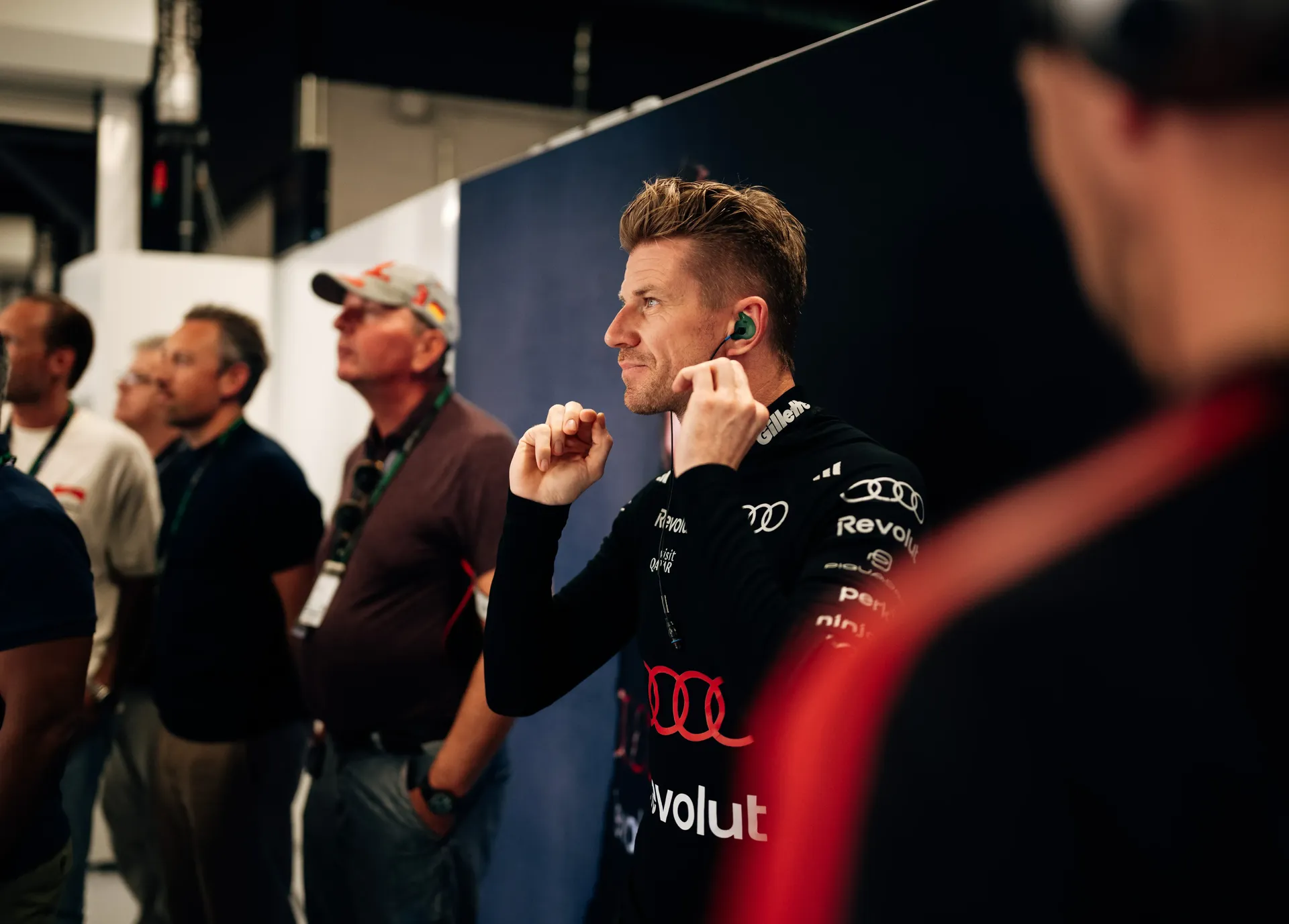 Nico Hulkenberg adjusting his earpiece while standing in the garage, with team members watching track action in the background.